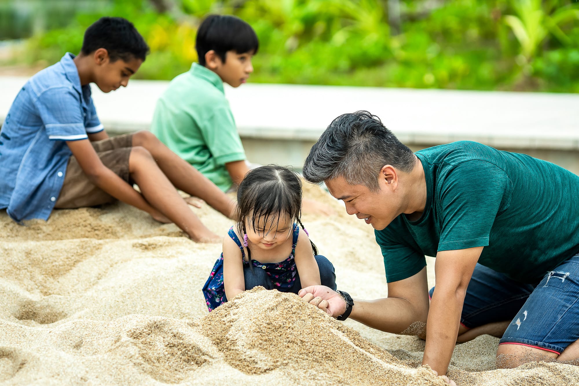 Several figures are sitting and playing in the sand outdoors, surrounded by greenery.