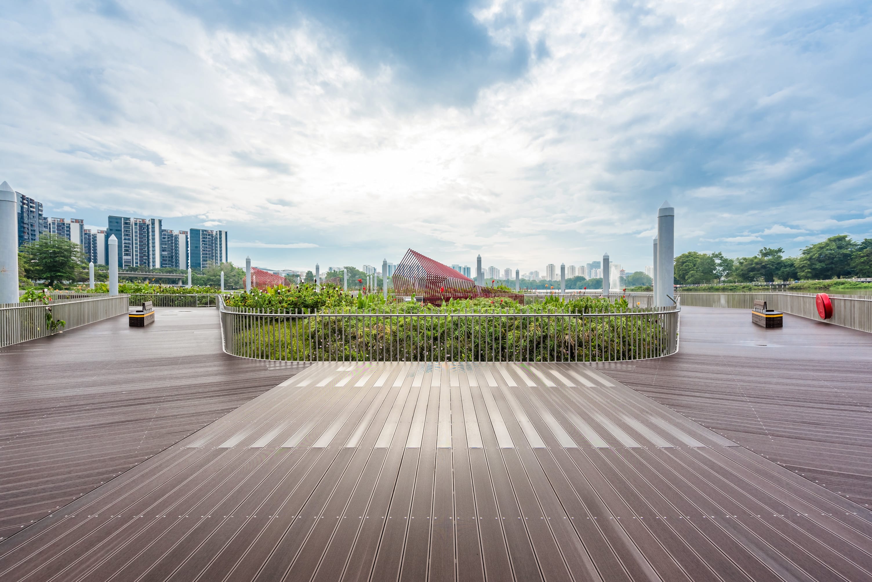 An open wooden deck with benches overlooking greenery and city buildings under a cloudy sky.