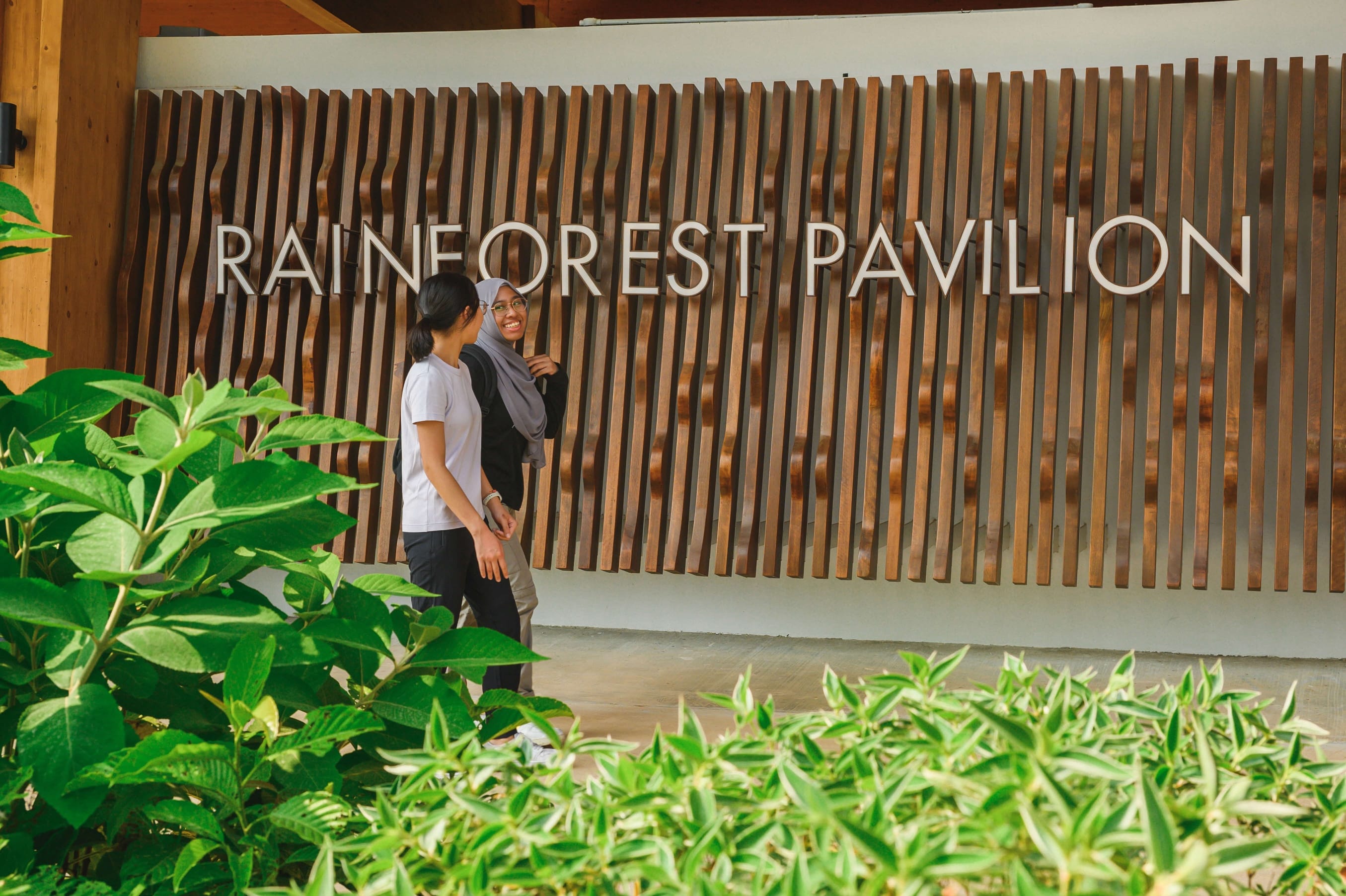 Two people stand in front of the "Rainforest Pavilion" wooden sign, surrounded by green foliage.