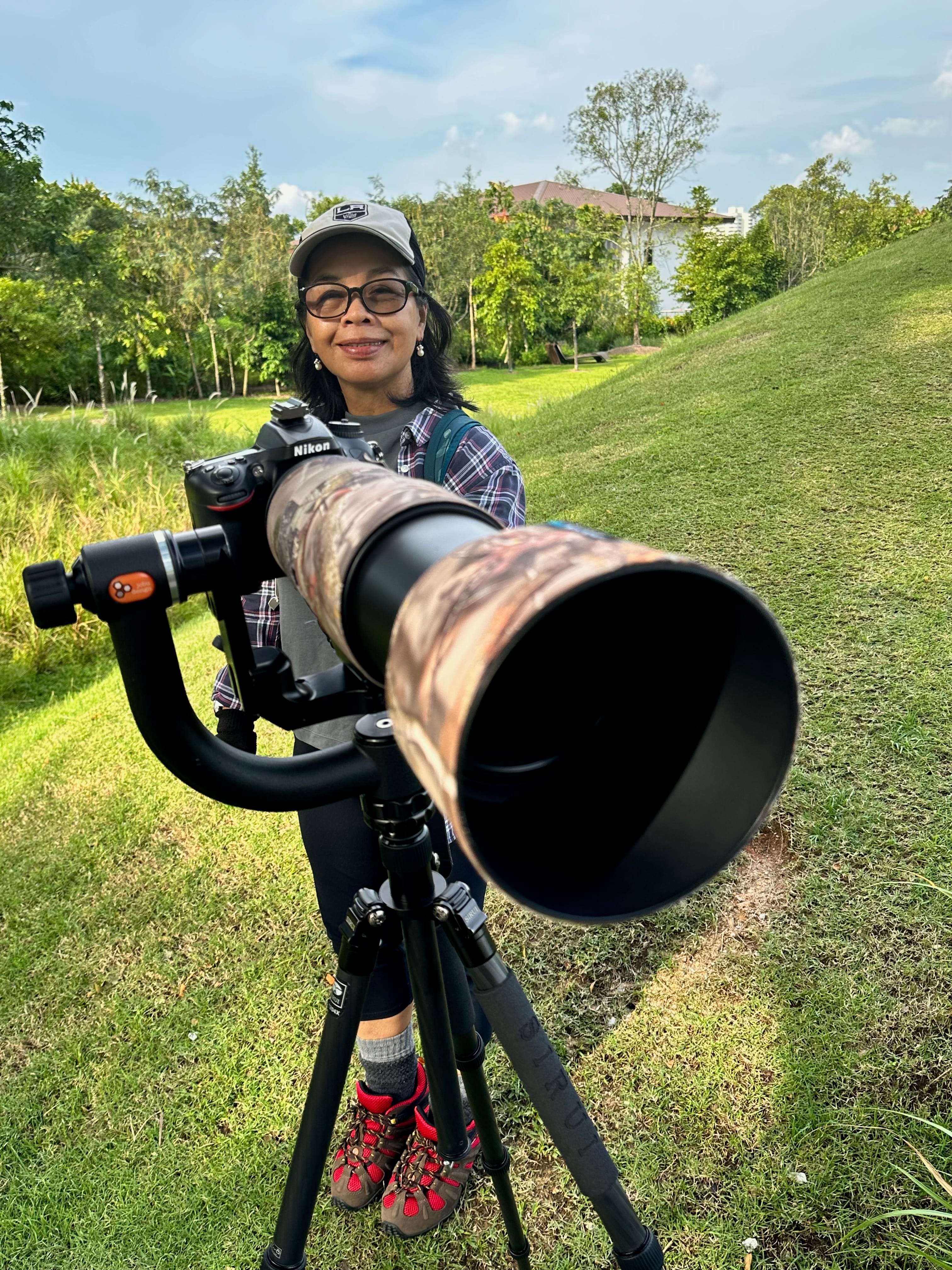 Person standing on grass with a Nikon camera and long lens on a tripod, wearing a plaid shirt and red hiking boots.