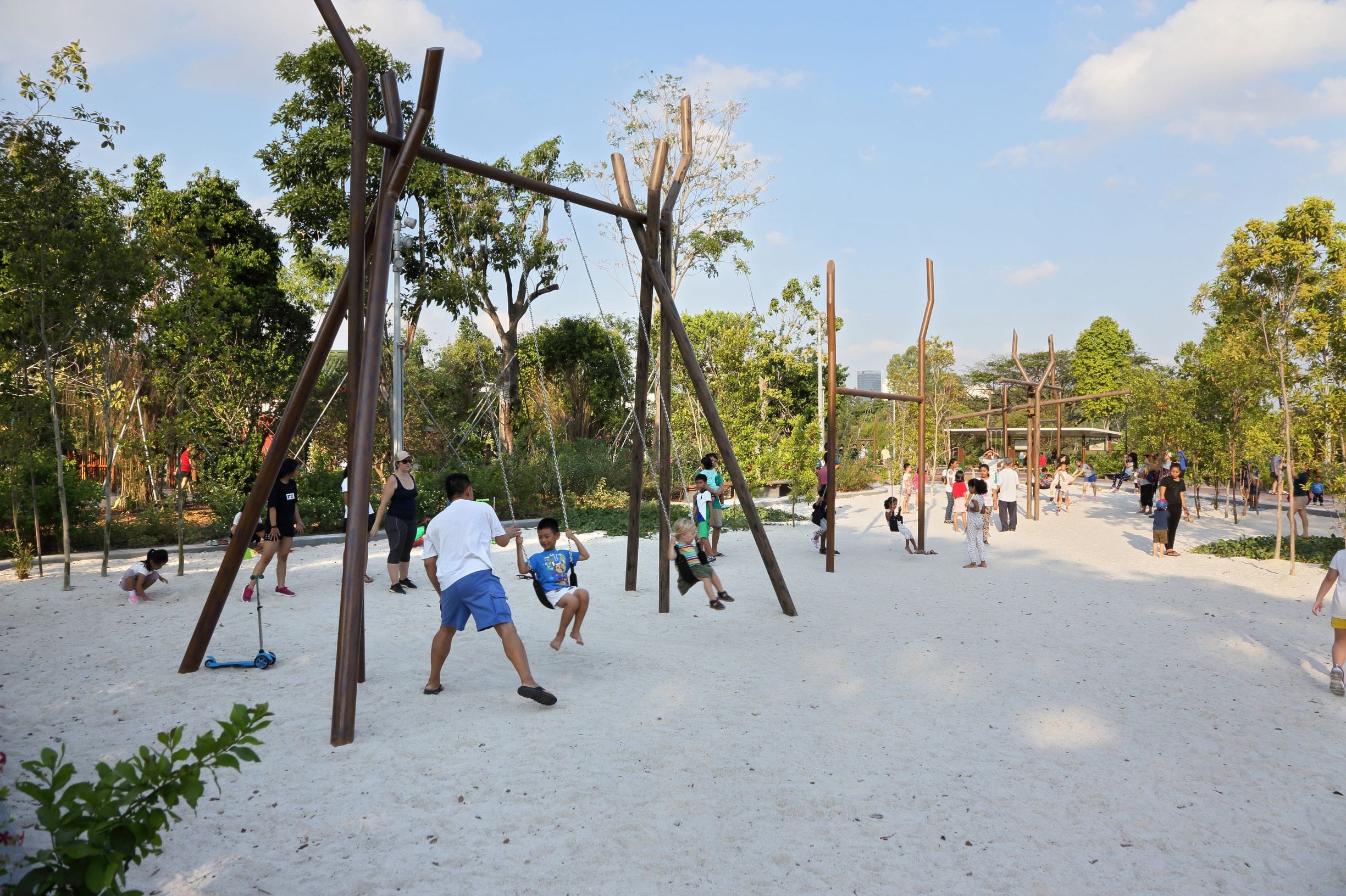 Children and adults enjoy swings in a park with sandy ground and many trees on a sunny day.