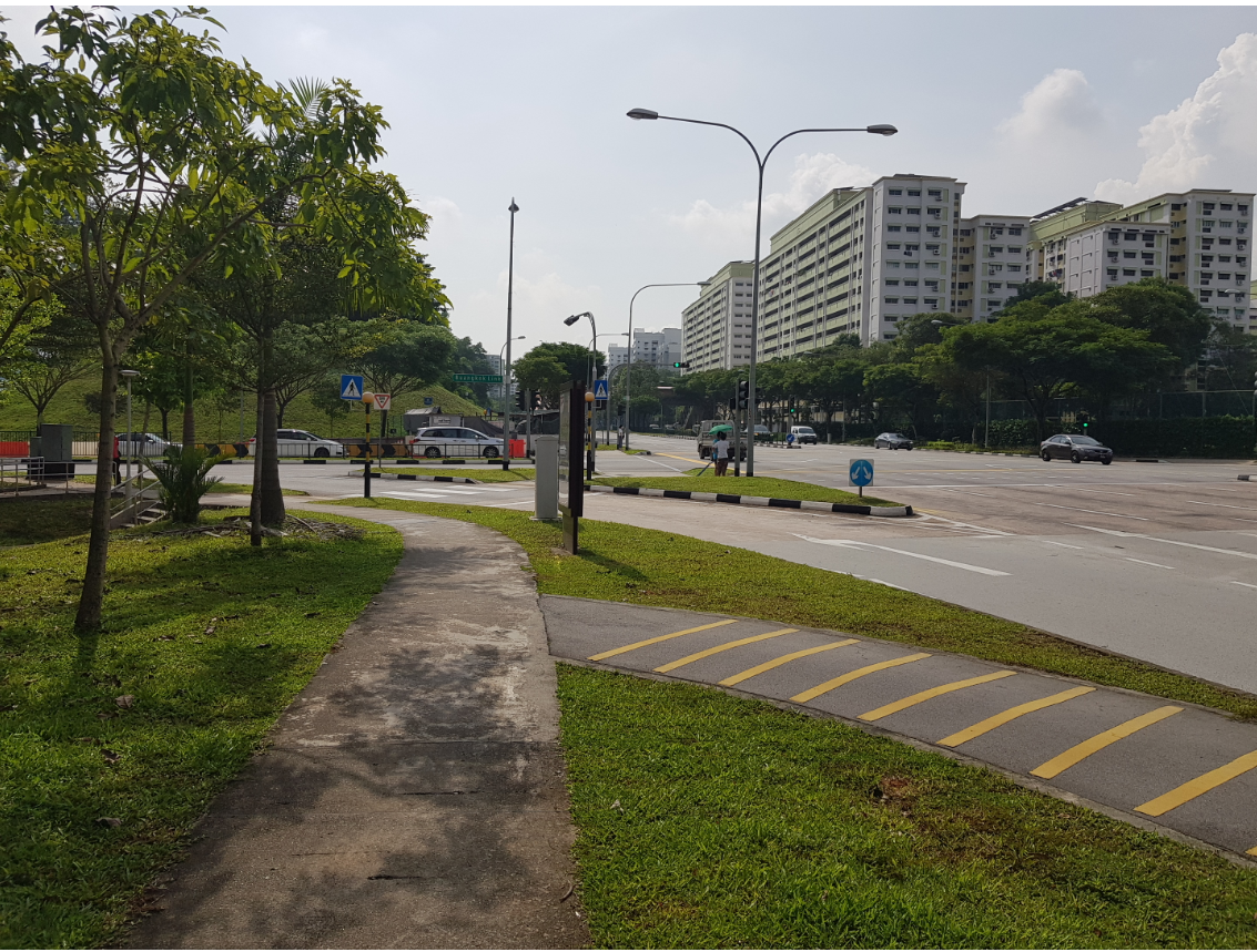 A sidewalk beside a road with cars, trees, and tall apartment buildings in the background under a cloudy sky.