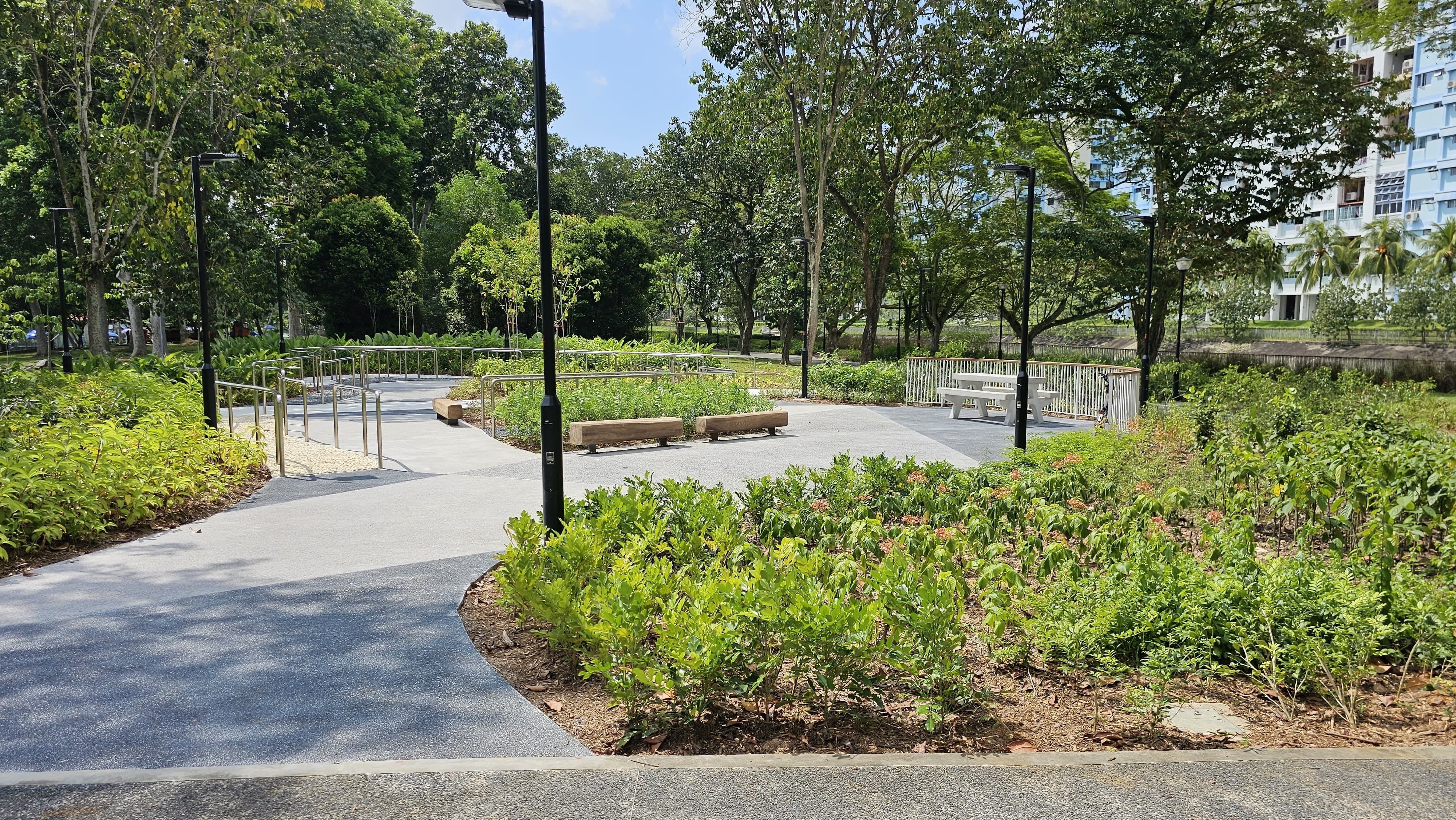 Curving paved path through a leafy park with railings, benches, lampposts, and an apartment block beyond trees.