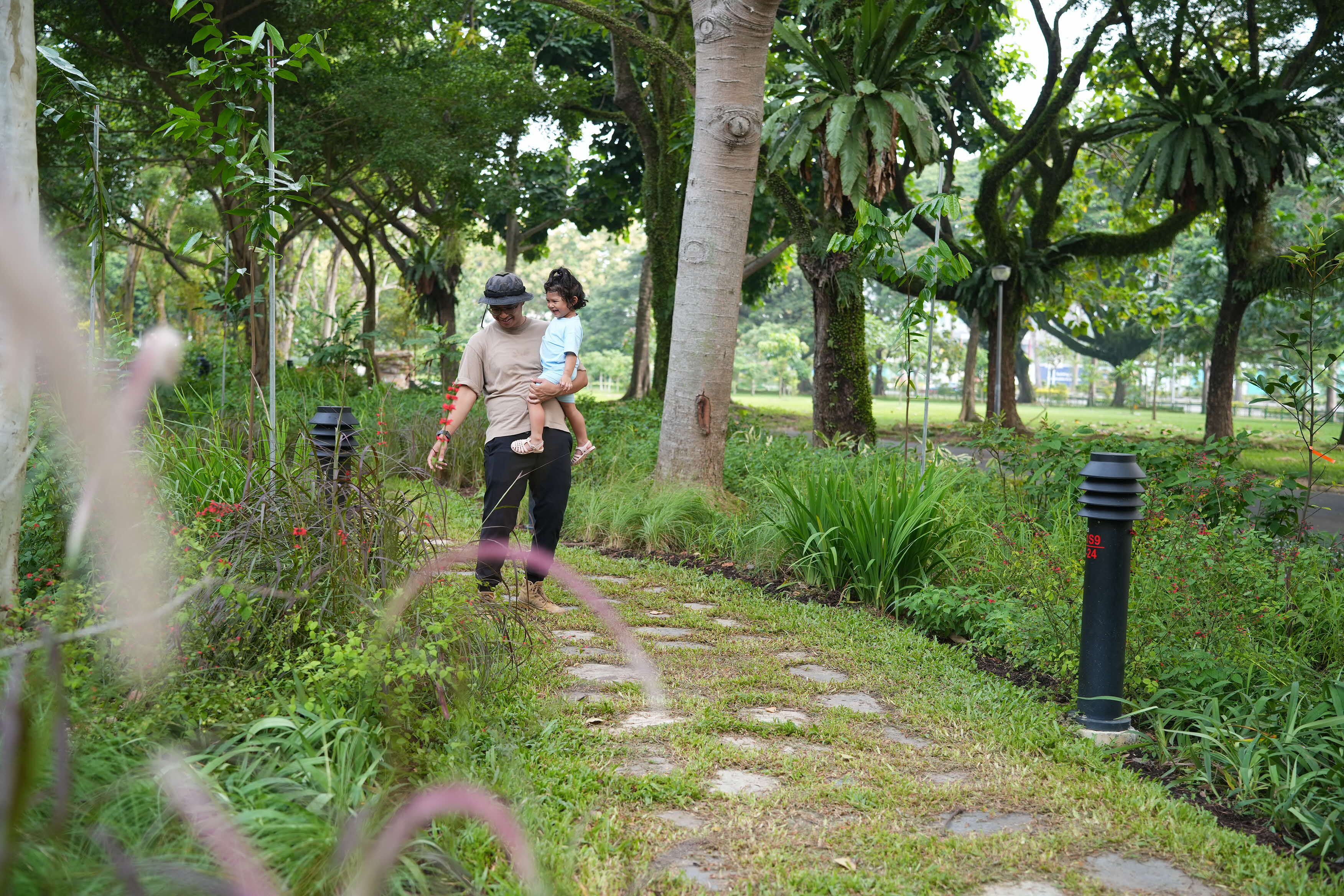 Man carries a child along a stone path through a lush park with tall trees, shrubs, and small lamp posts.