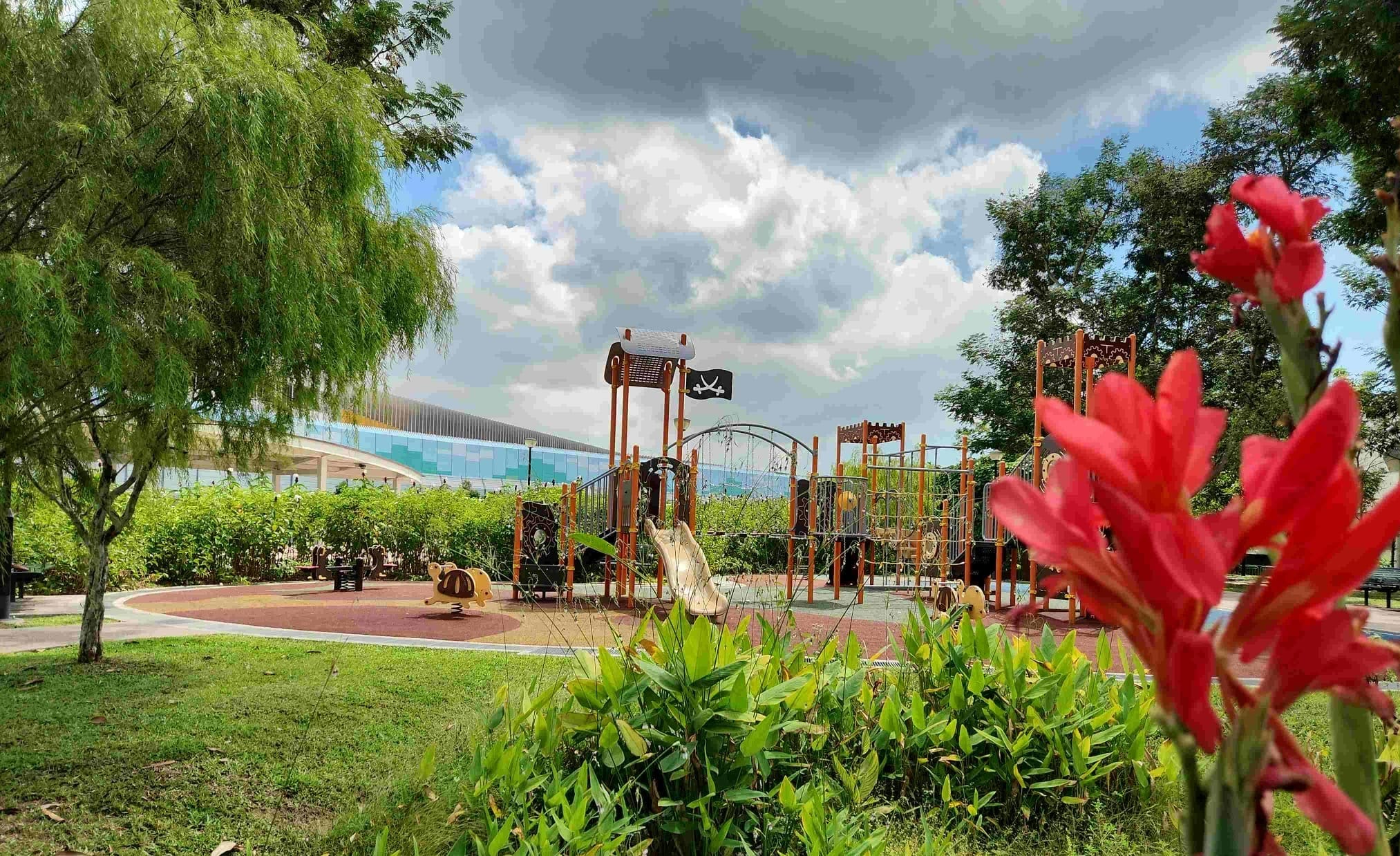 Playground with slides and climbing frames under cloudy sky, trees around, red flowers in foreground.