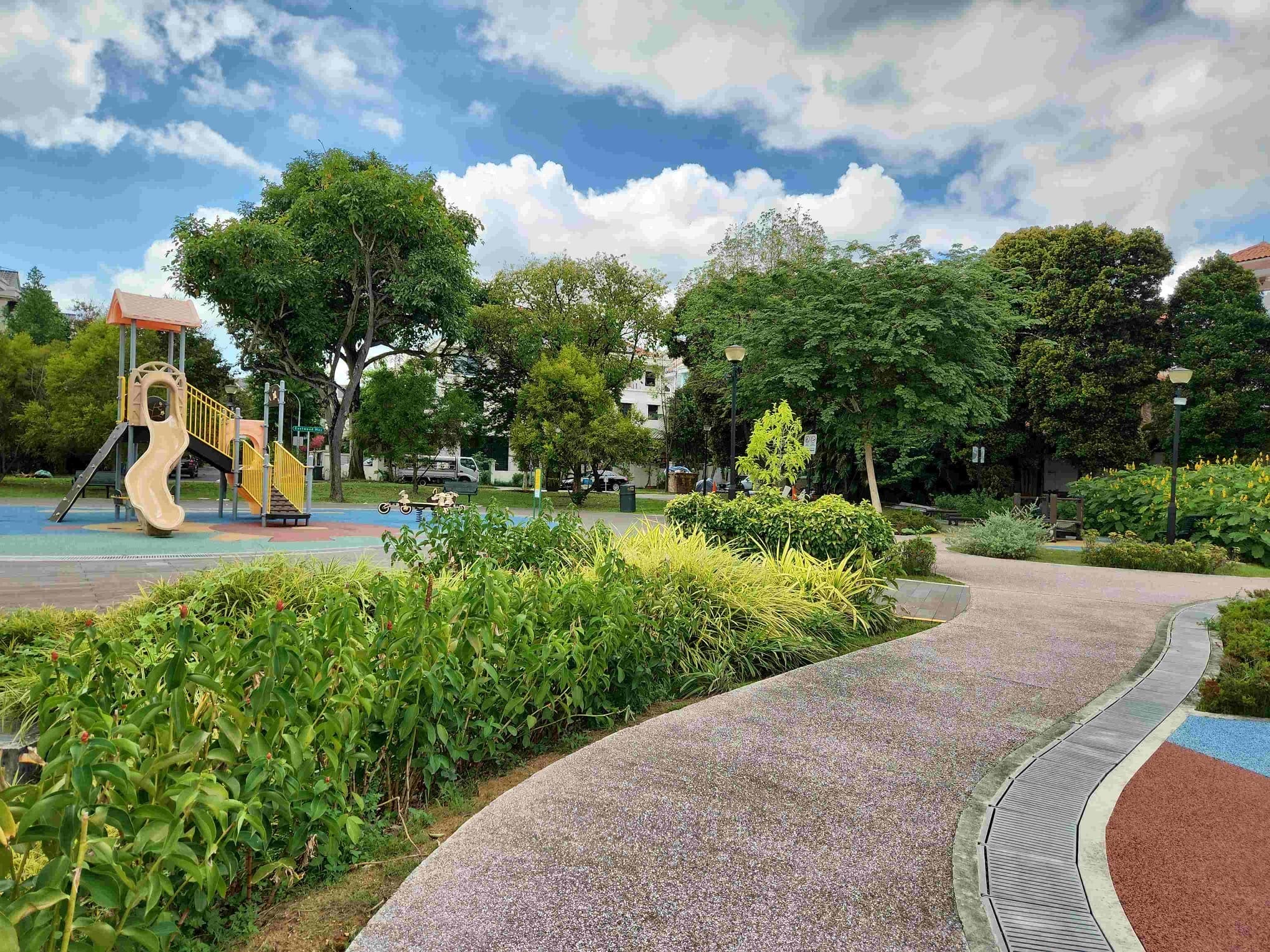 Curving park path beside green shrubs, with a playground slide, tall trees, and a cloudy blue sky.