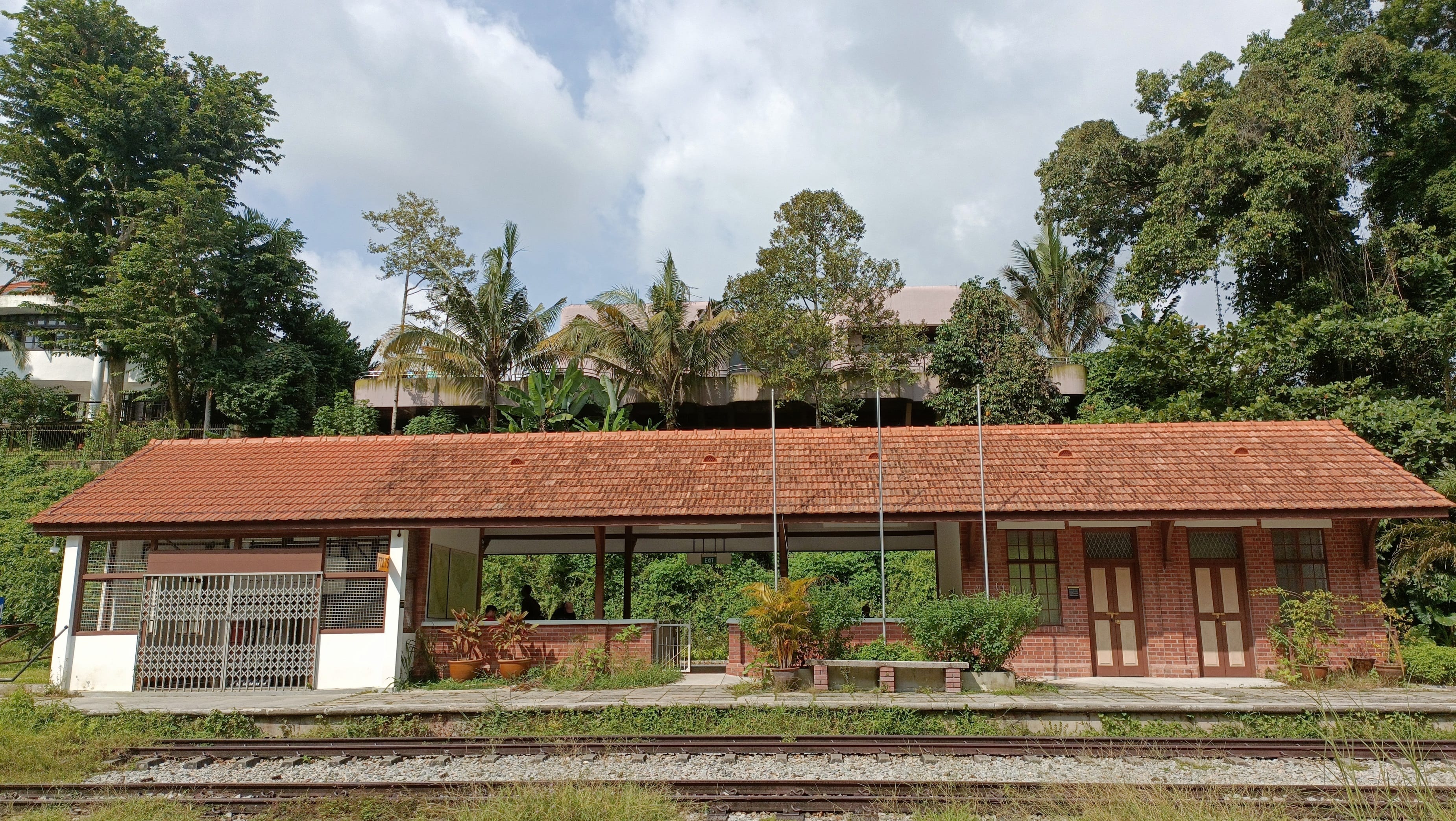 A small brick train station with a tiled roof, surrounded by lush trees and a building in the background.