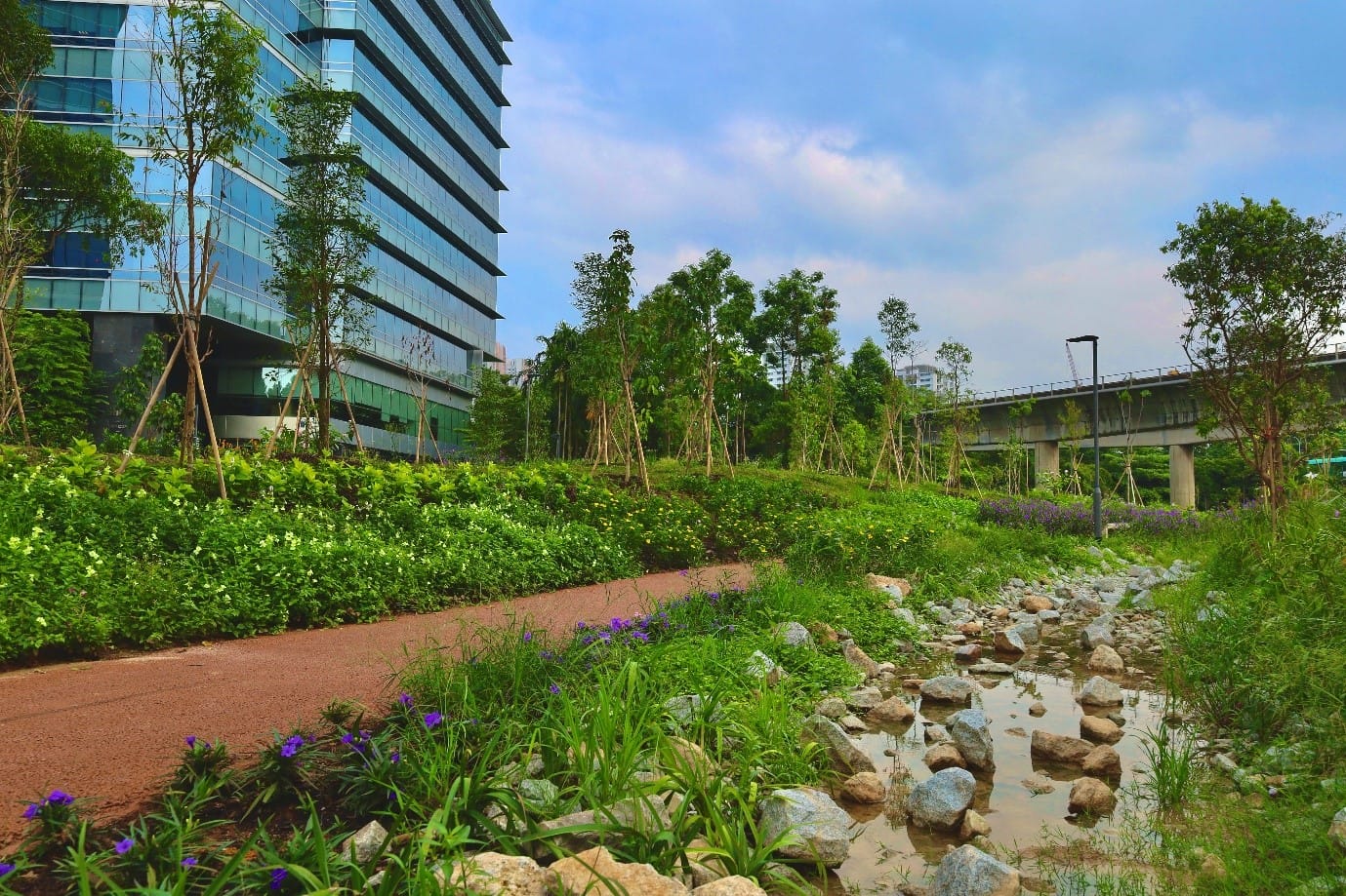 A modern glass building with a green garden, a walkway, rocks, and a small stream, under a partly cloudy sky.