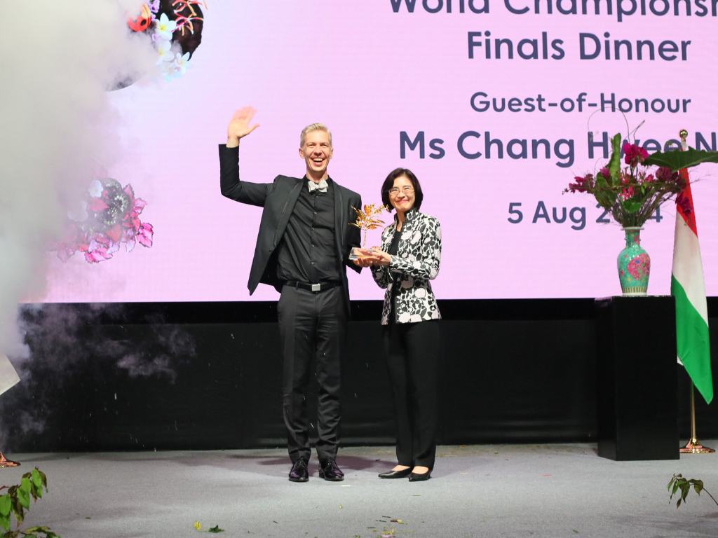 Two people are standing on a stage, one waving, the other holding a floral trophy, with event details displayed behind them.