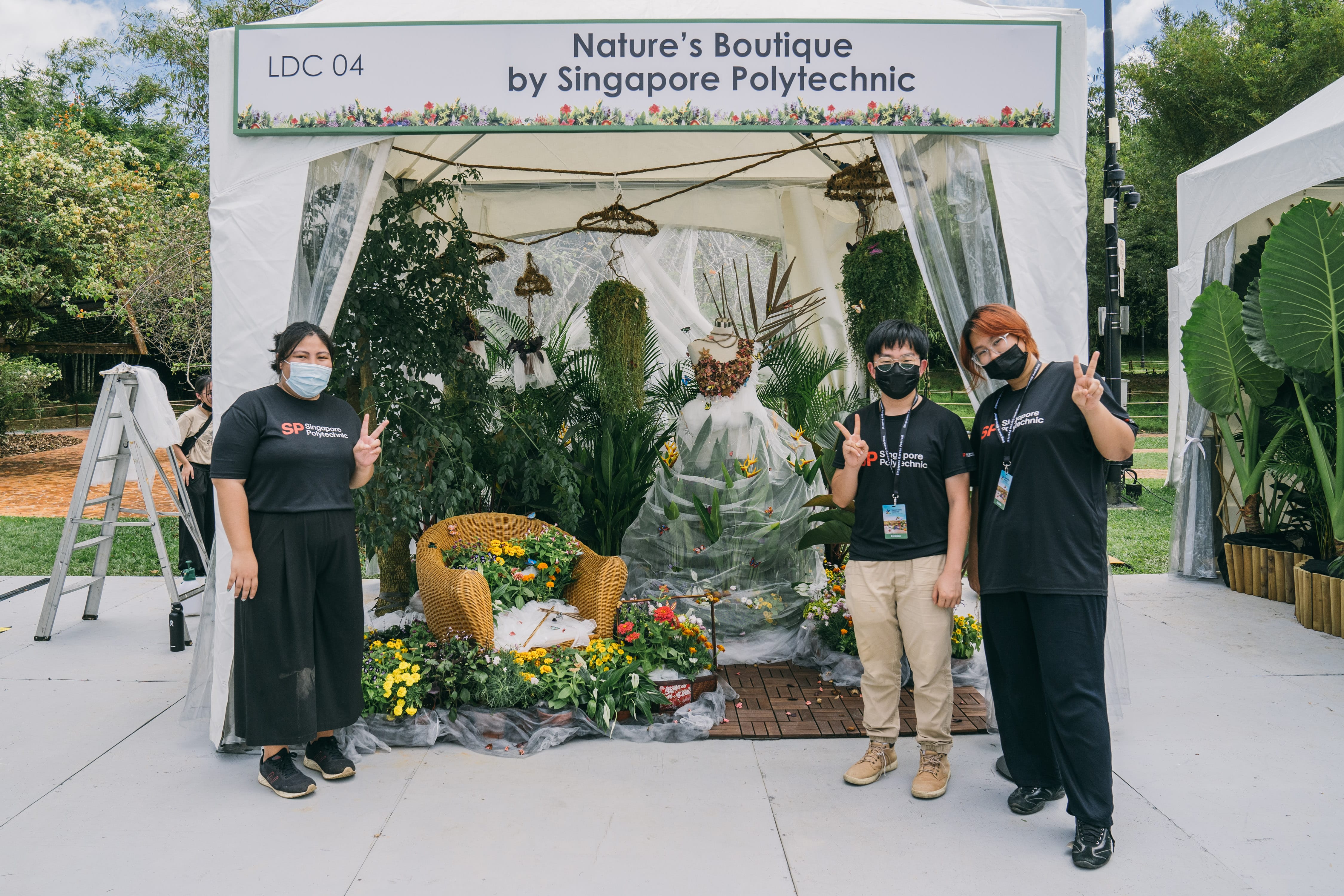 Three people stand in front of a decorated outdoor booth with plants and a sign reading "Nature's Boutique by Singapore Polytechnic."