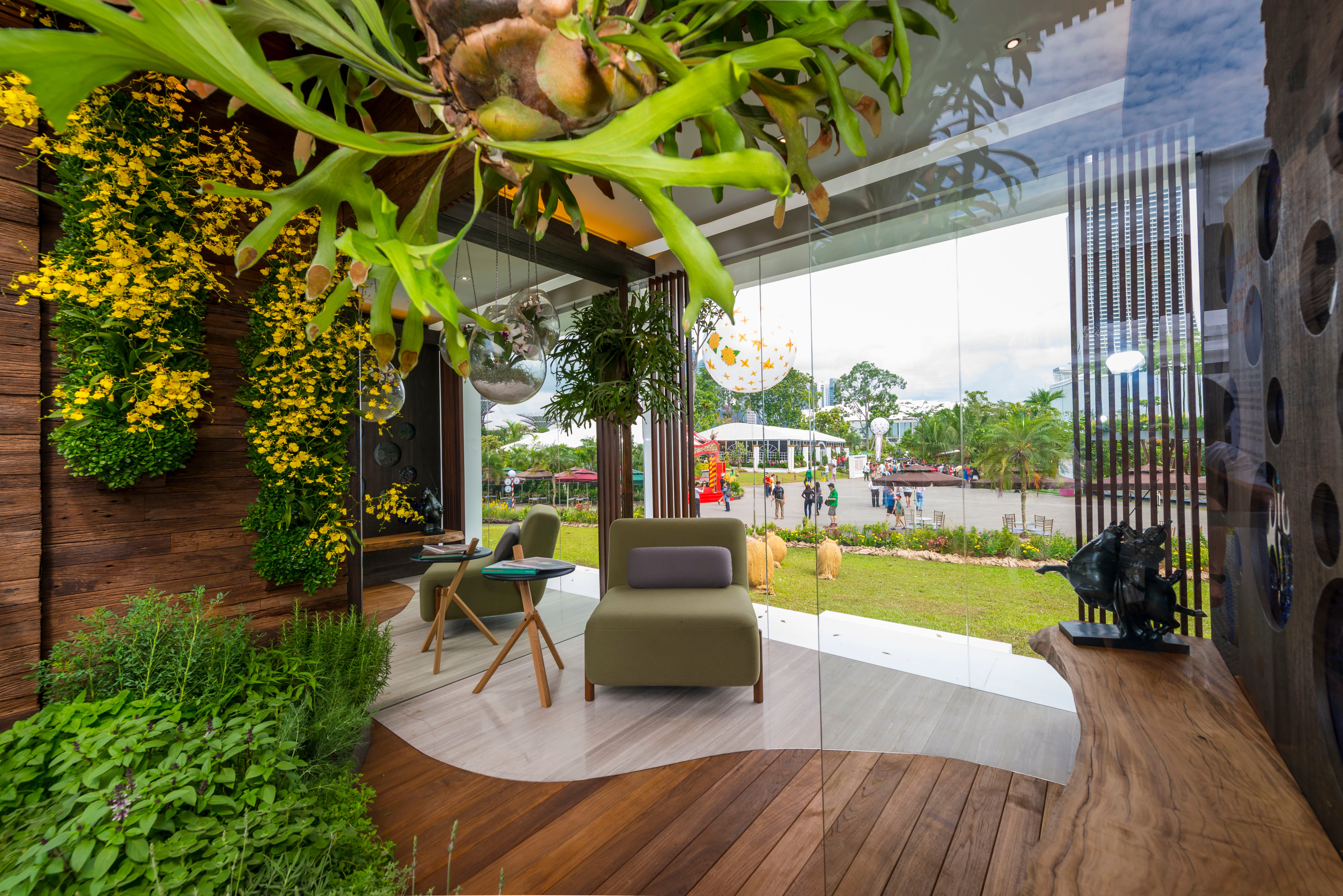 A modern outdoor seating area with green plants, yellow flowers, and a view of people in the background through a glass wall.