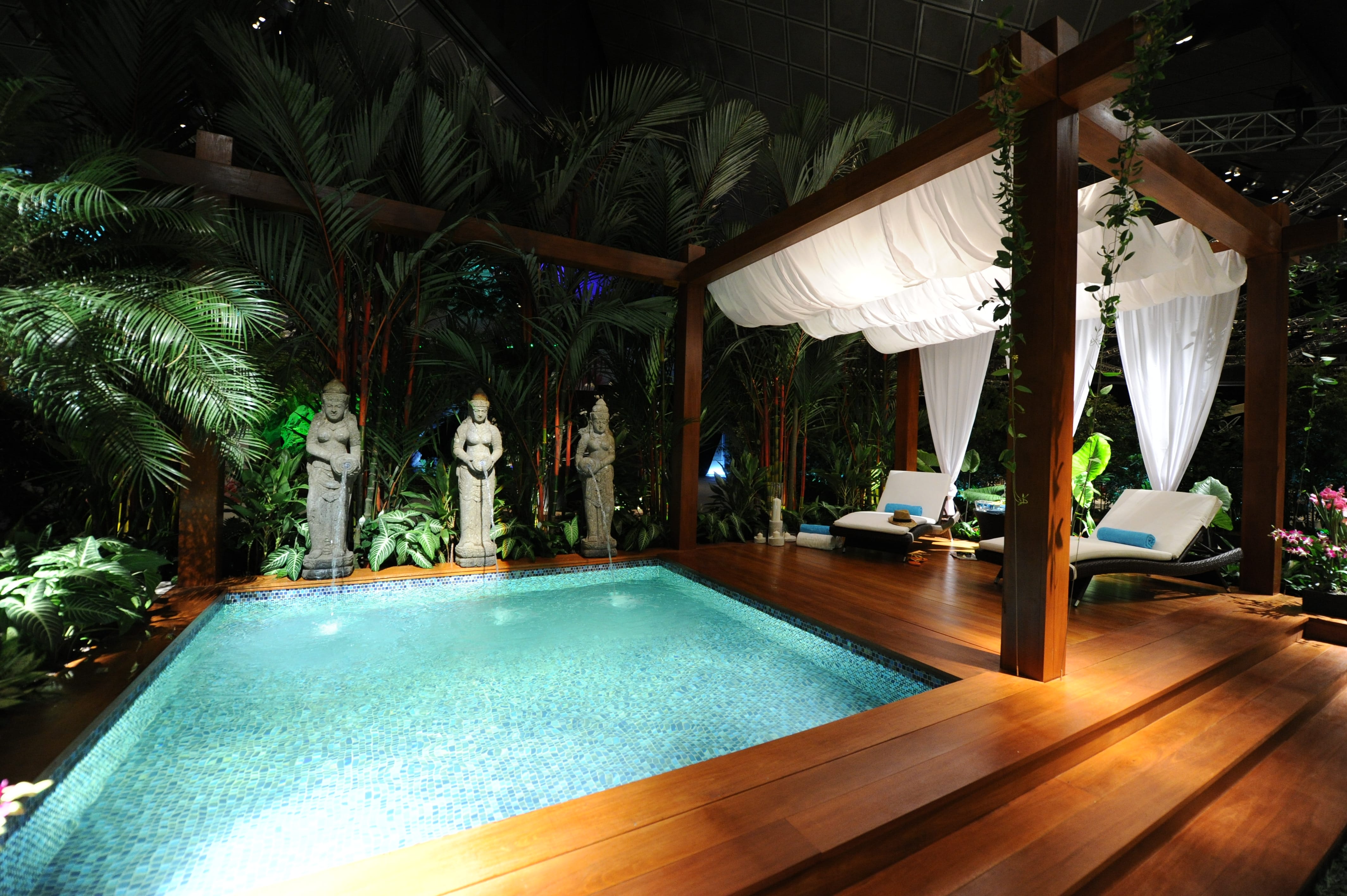 A luxurious outdoor pool surrounded by tropical plants, stone statues, and wooden deck with two lounge chairs under a canopy.