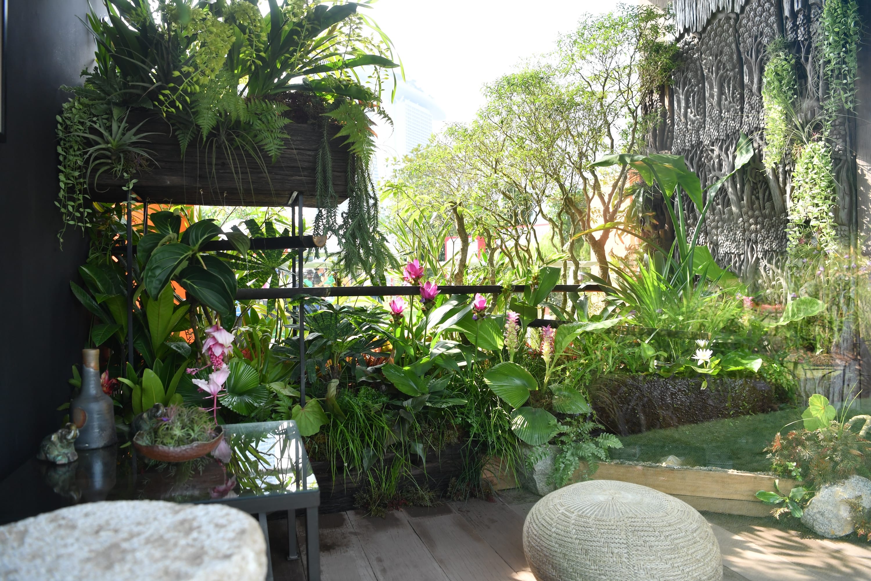 A lush indoor garden with diverse green plants, pink flowers, and a woven round ottoman.
