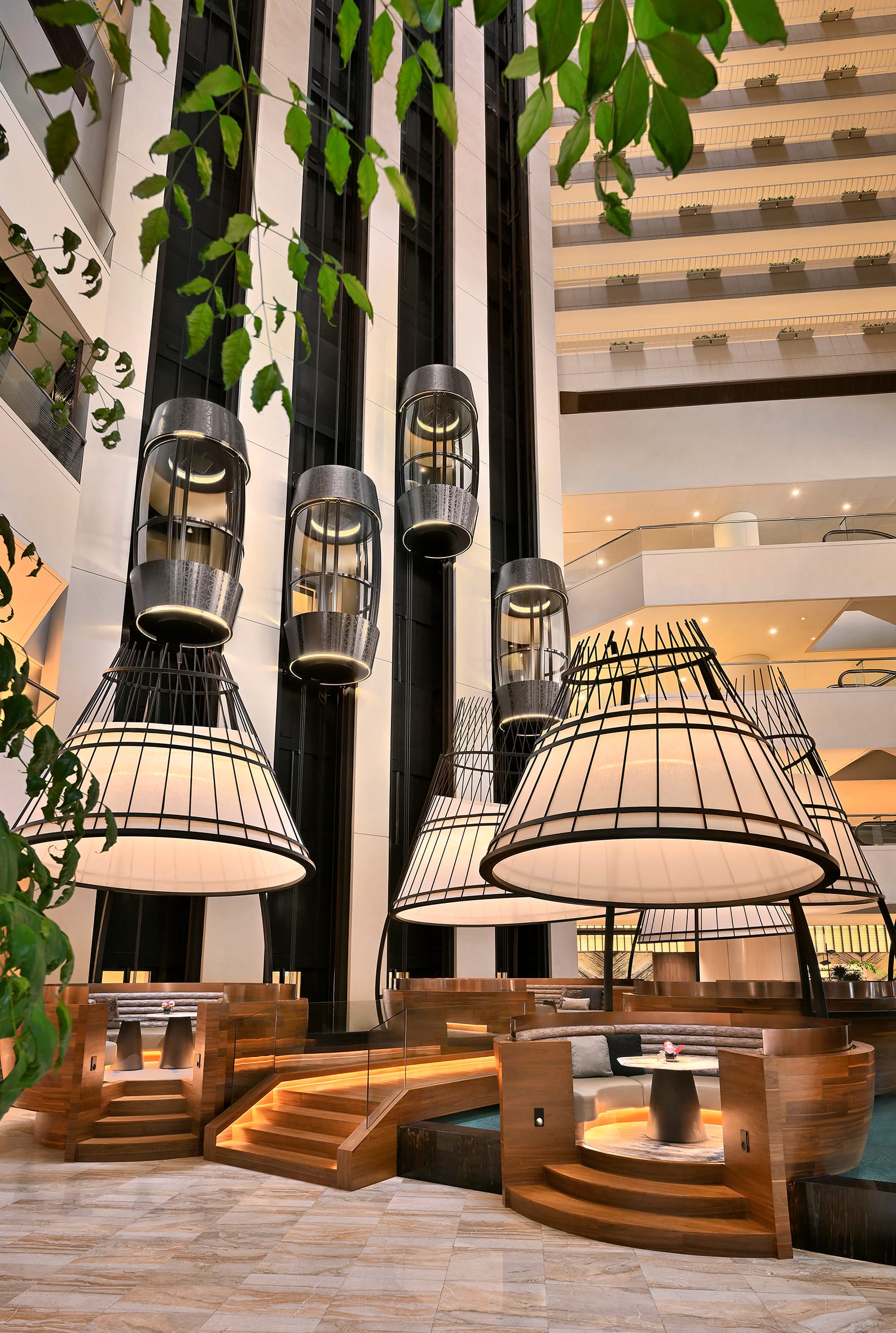 Luxurious hotel lobby with hanging lanterns, modern elevators, wooden seating pods, and decorative plants.
