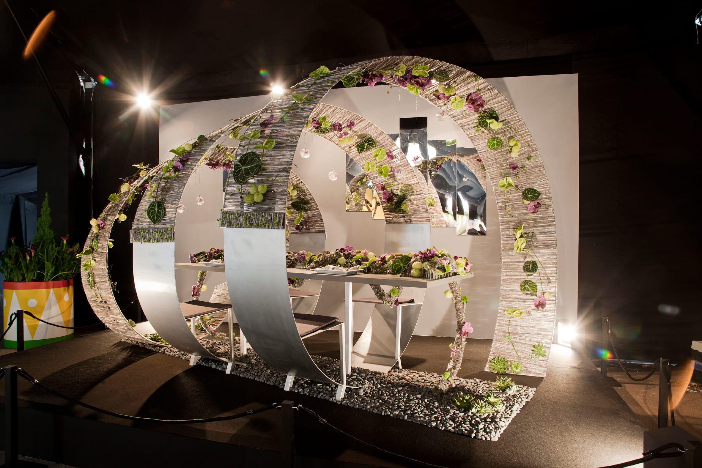 A table setting surrounded by decorative arches adorned with flowers and greenery, illuminated by spotlights.