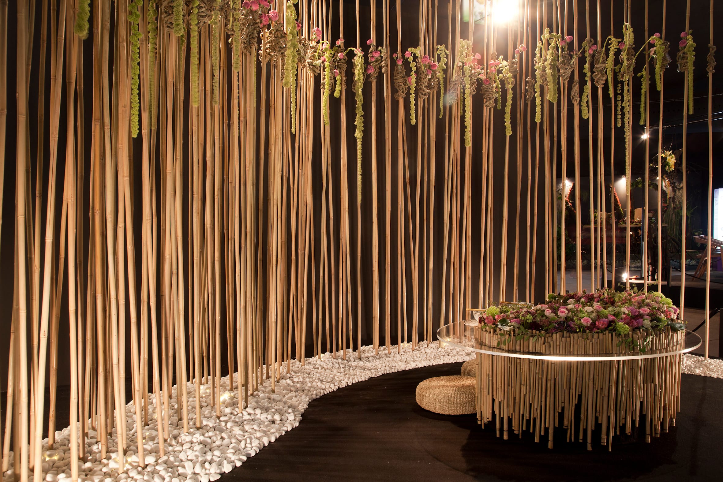 Tall bamboo sticks surrounded by white pebbles and hanging green and pink flowers in a dark lit room with a circular table.