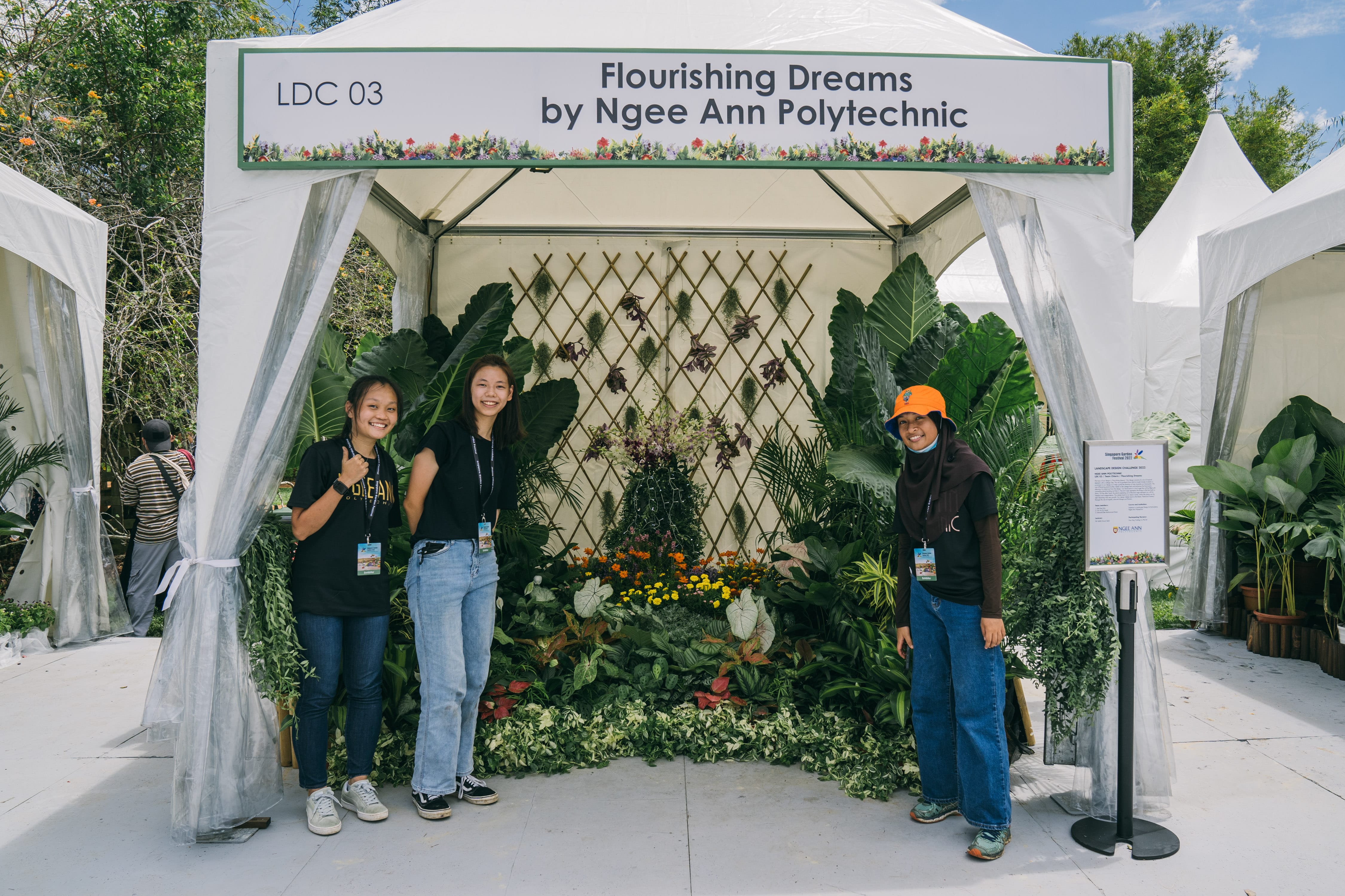 A garden display under a tent, with plants arranged around a trellis backdrop, and three people standing nearby.
