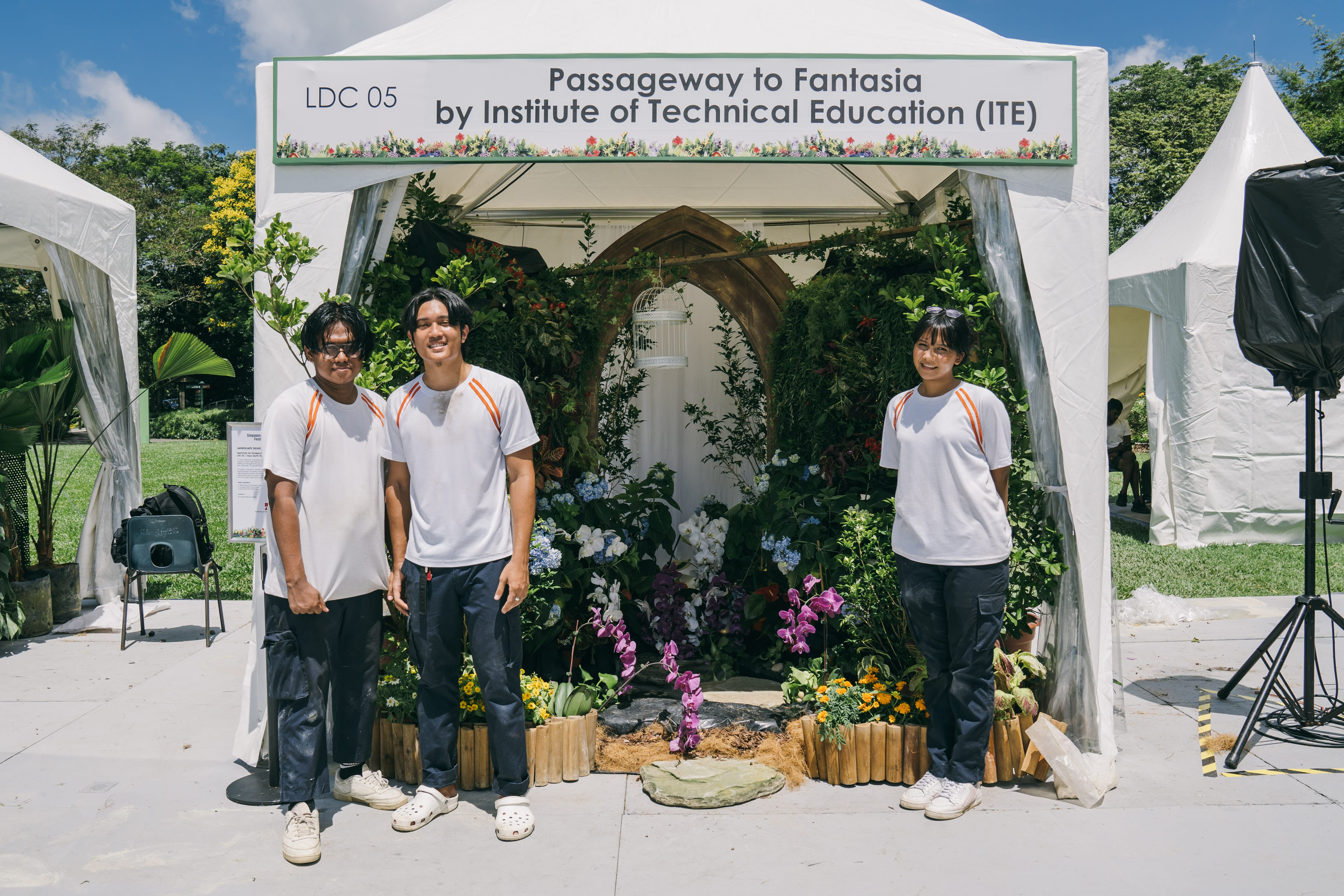 Three people wearing white shirts stand in front of a flower-decorated tent labeled "Passageway to Fantasia by Institute of Technical Education."