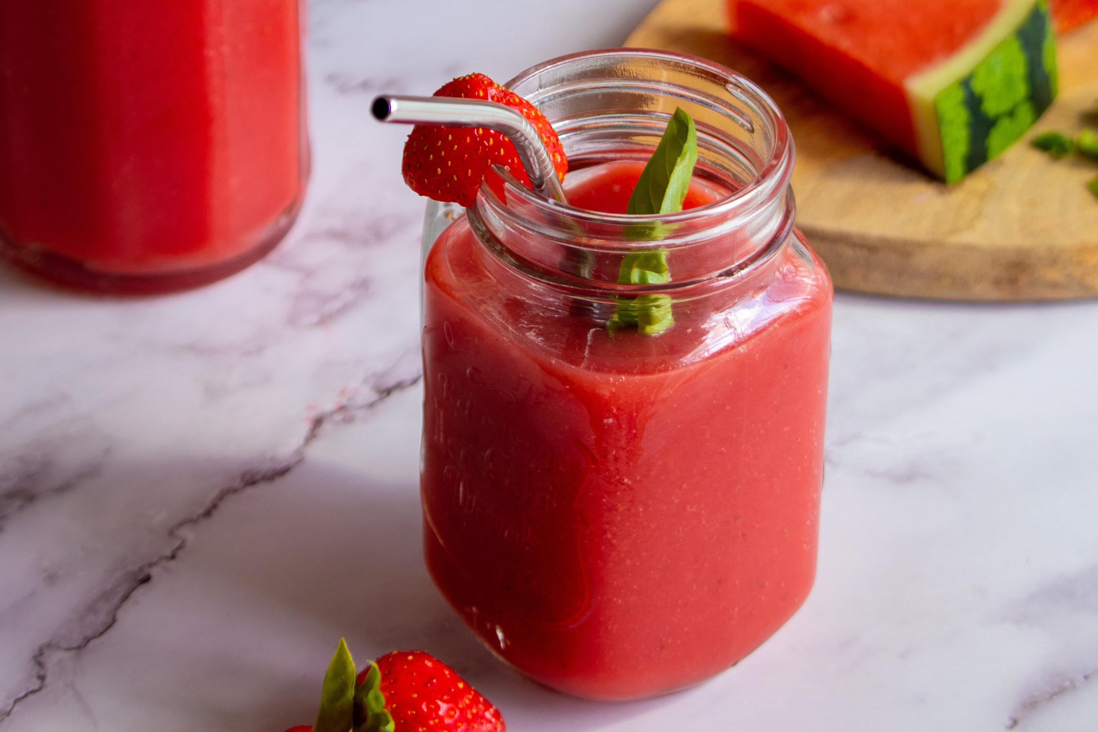 Glass jar with red smoothie, garnished with a strawberry slice, basil leaves, and a metal straw, on a marble surface.