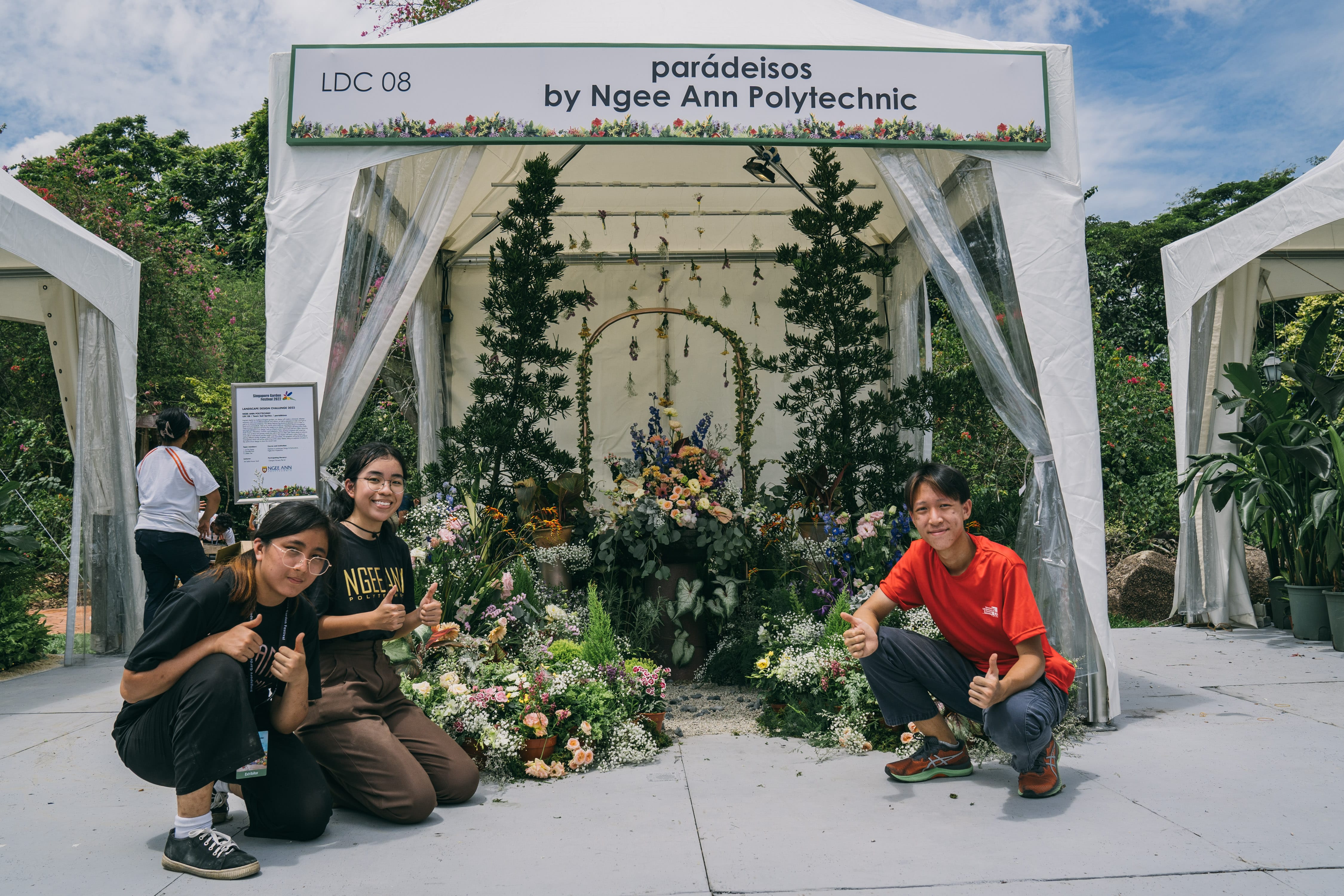 An outdoor floral display with lush greenery, vibrant flowers, and a banner reading "parádeisos by Ngee Ann Polytechnic."