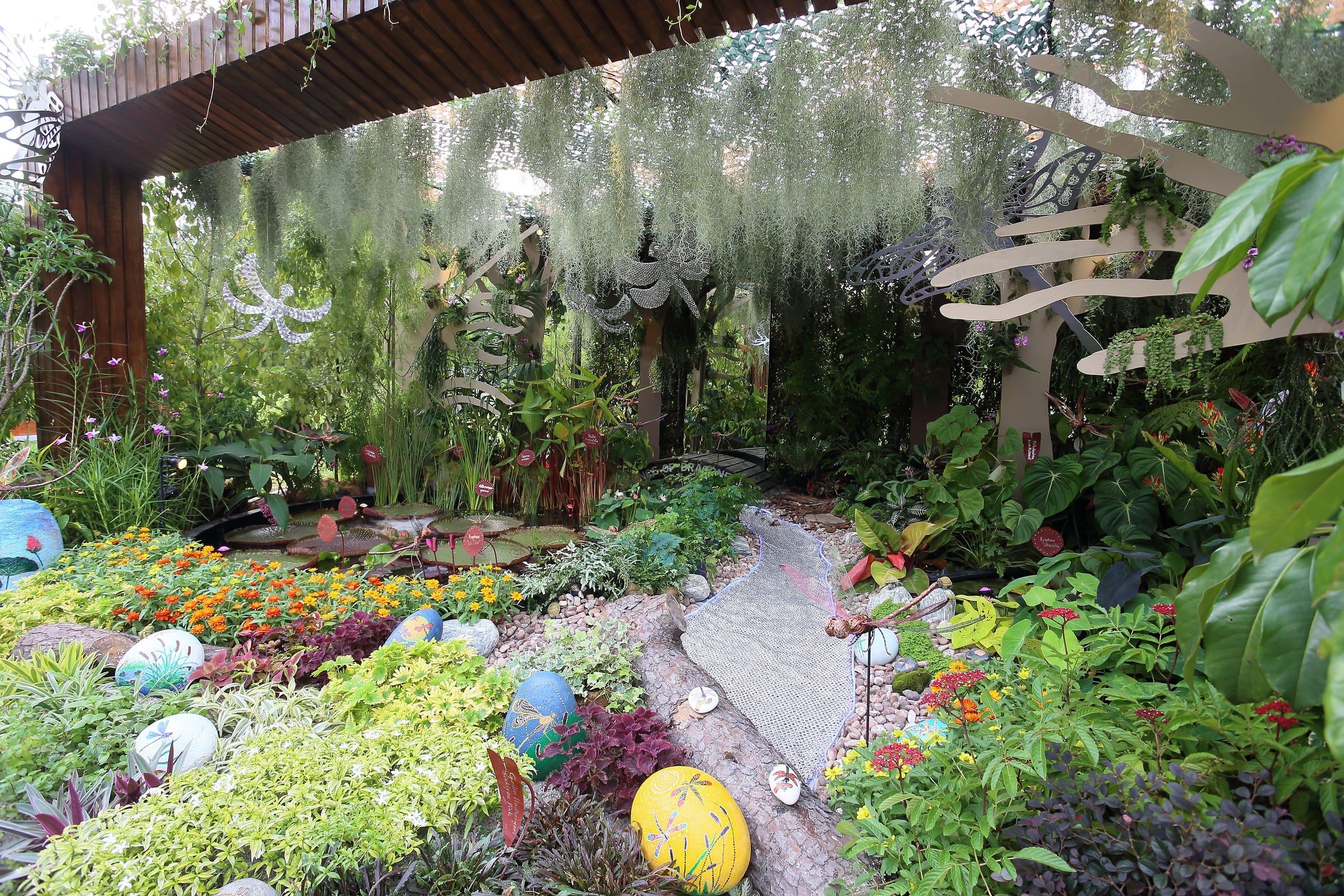 A colorful garden under a wooden pergola, featuring plants, flowers, hanging moss, and decorative dragonfly structures.