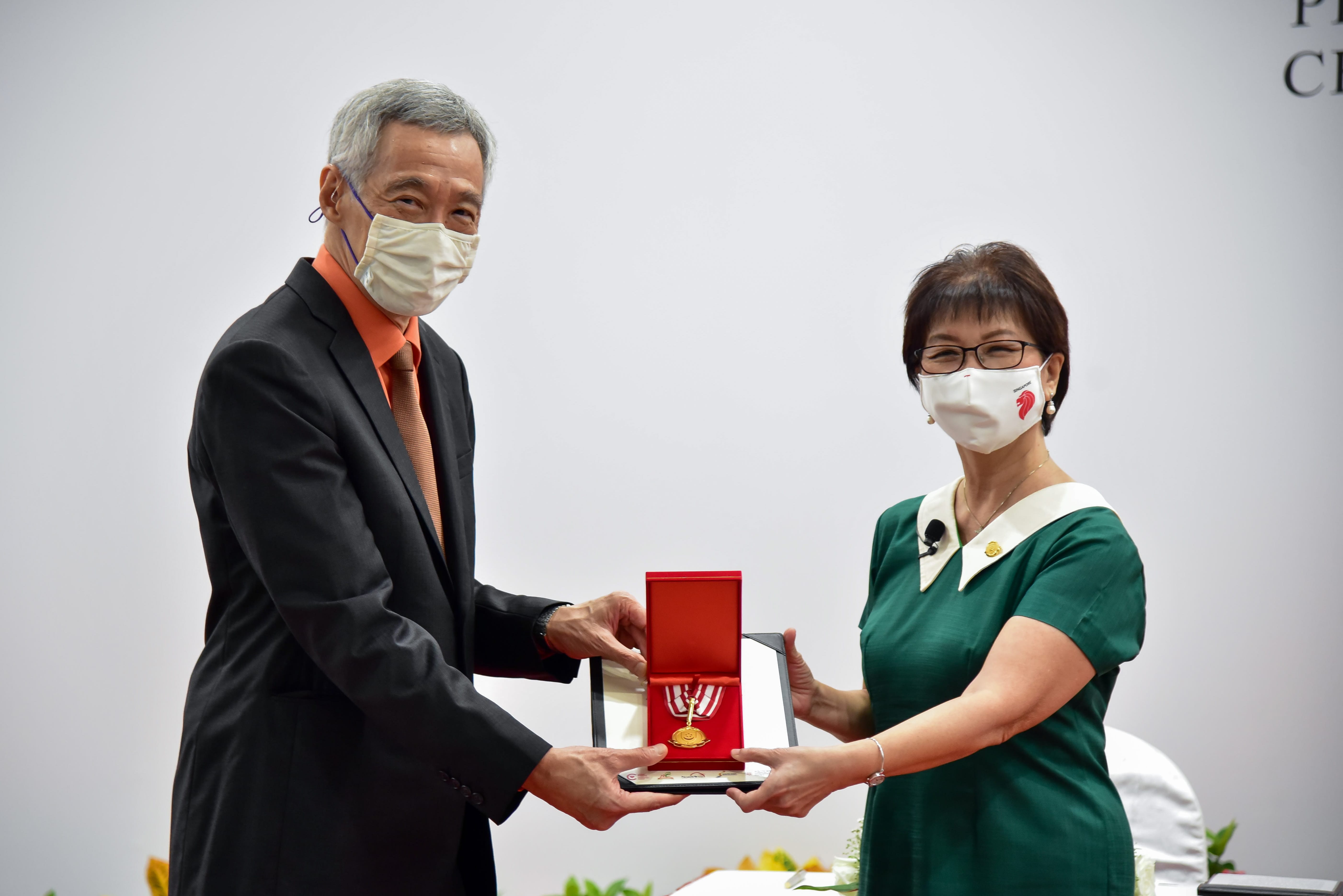 Two people exchange a red and gold medal in a box; one wears a green dress and the other a black suit with an orange shirt.