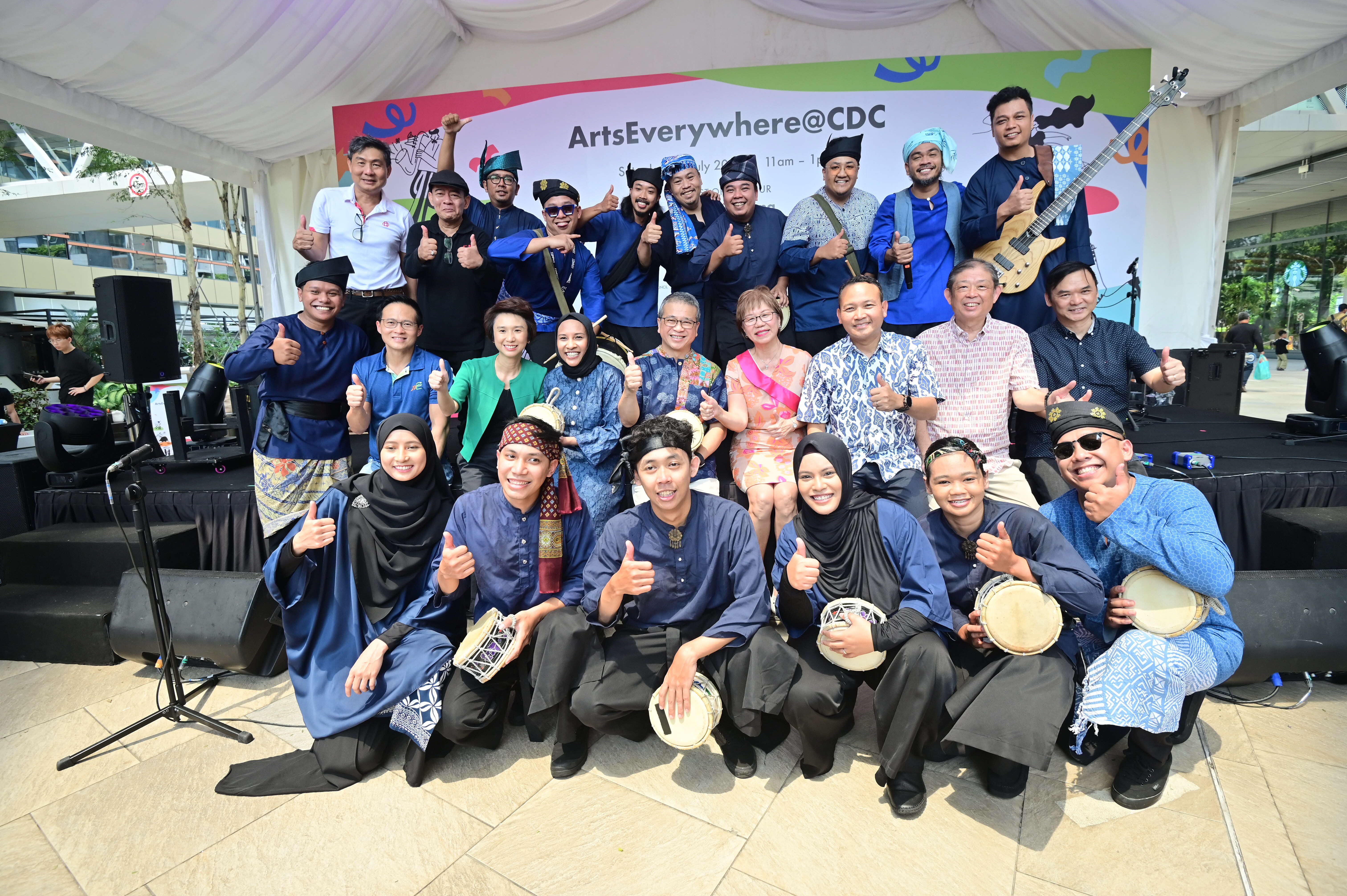A group of people in traditional attire and casual clothes pose with musical instruments and thumbs up under a canopy.