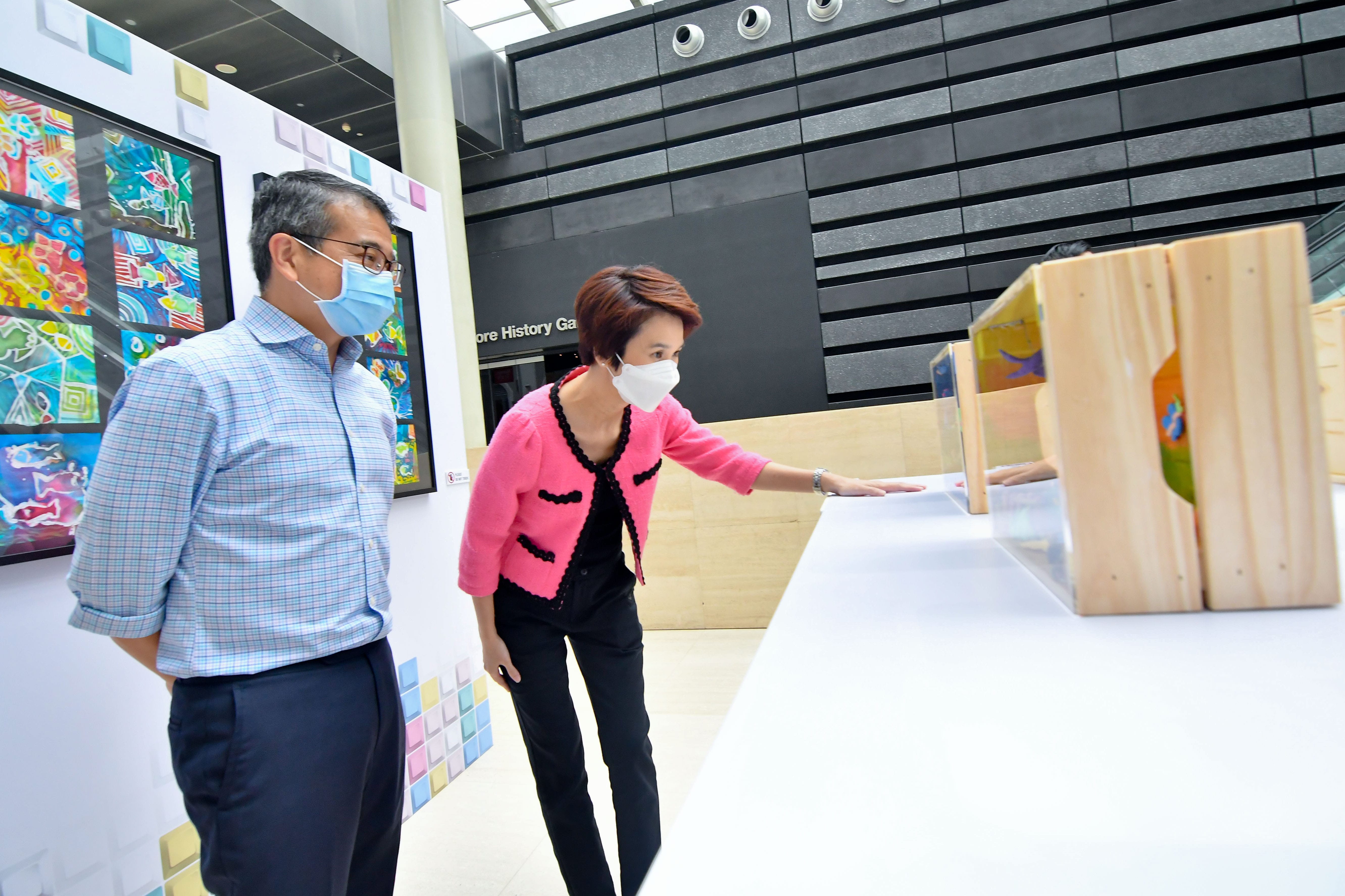 Two people stand near colorful artwork on display in a modern gallery with light wood and dark panelled walls.