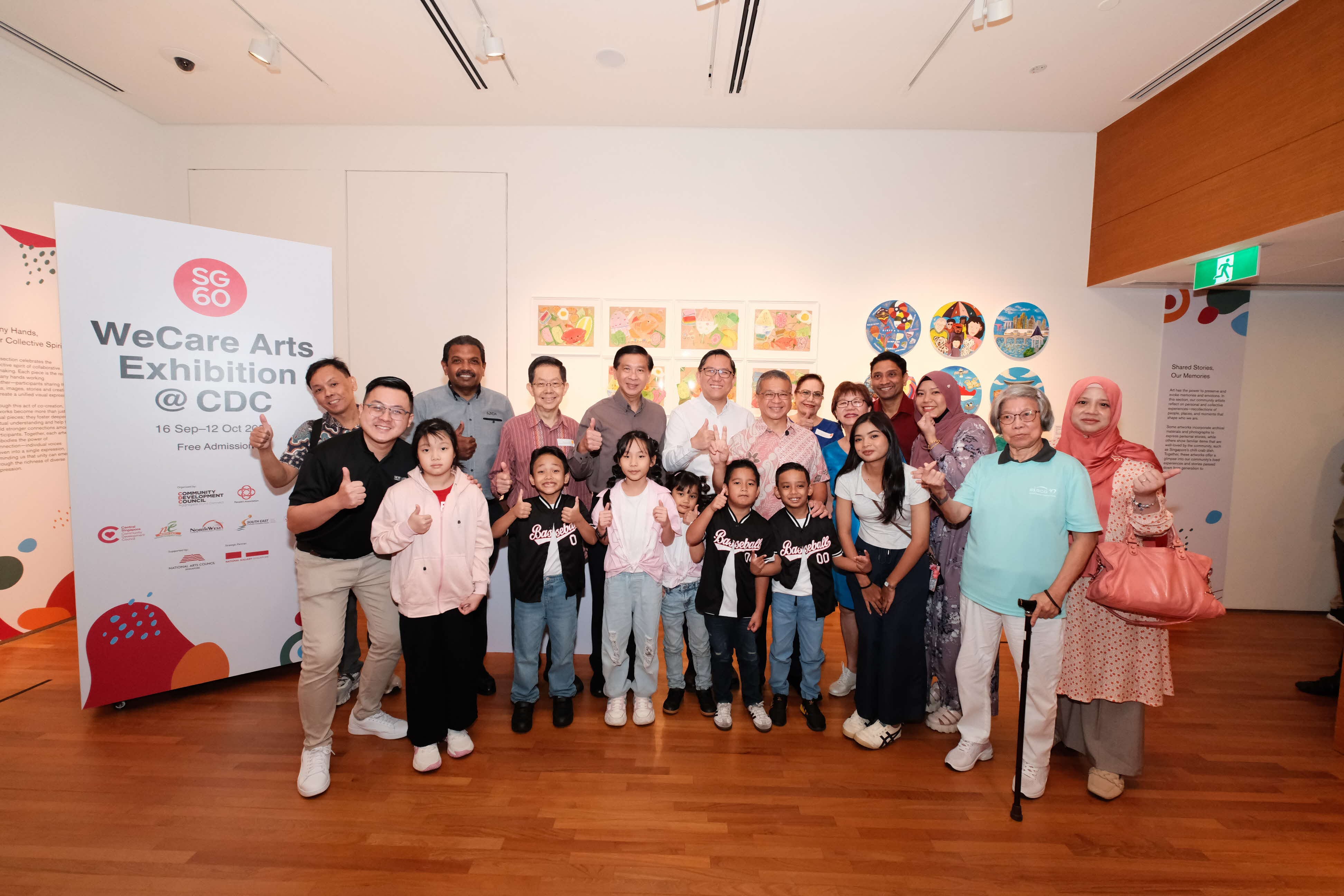 A group of people pose together at an arts exhibition, standing on a wooden floor next to a large signboard.