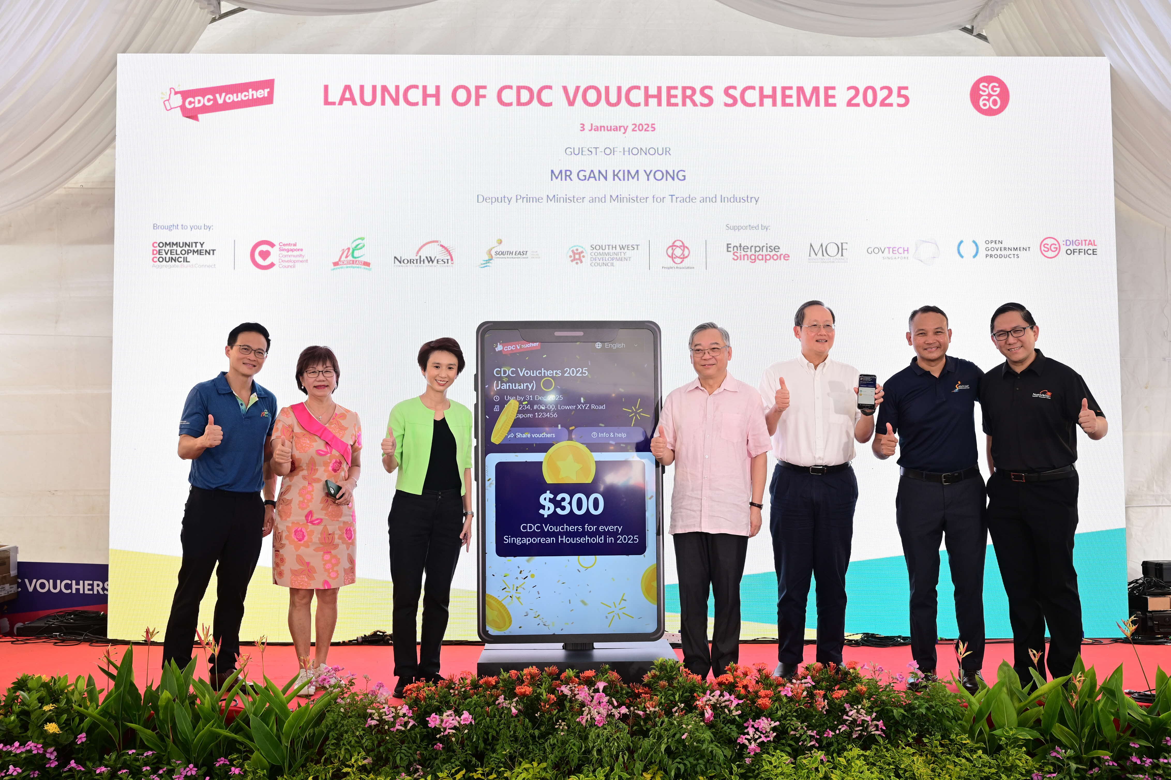 Six people standing on stage with a large phone screen displaying "$300 CDC Vouchers for every Singaporean Household in 2025".