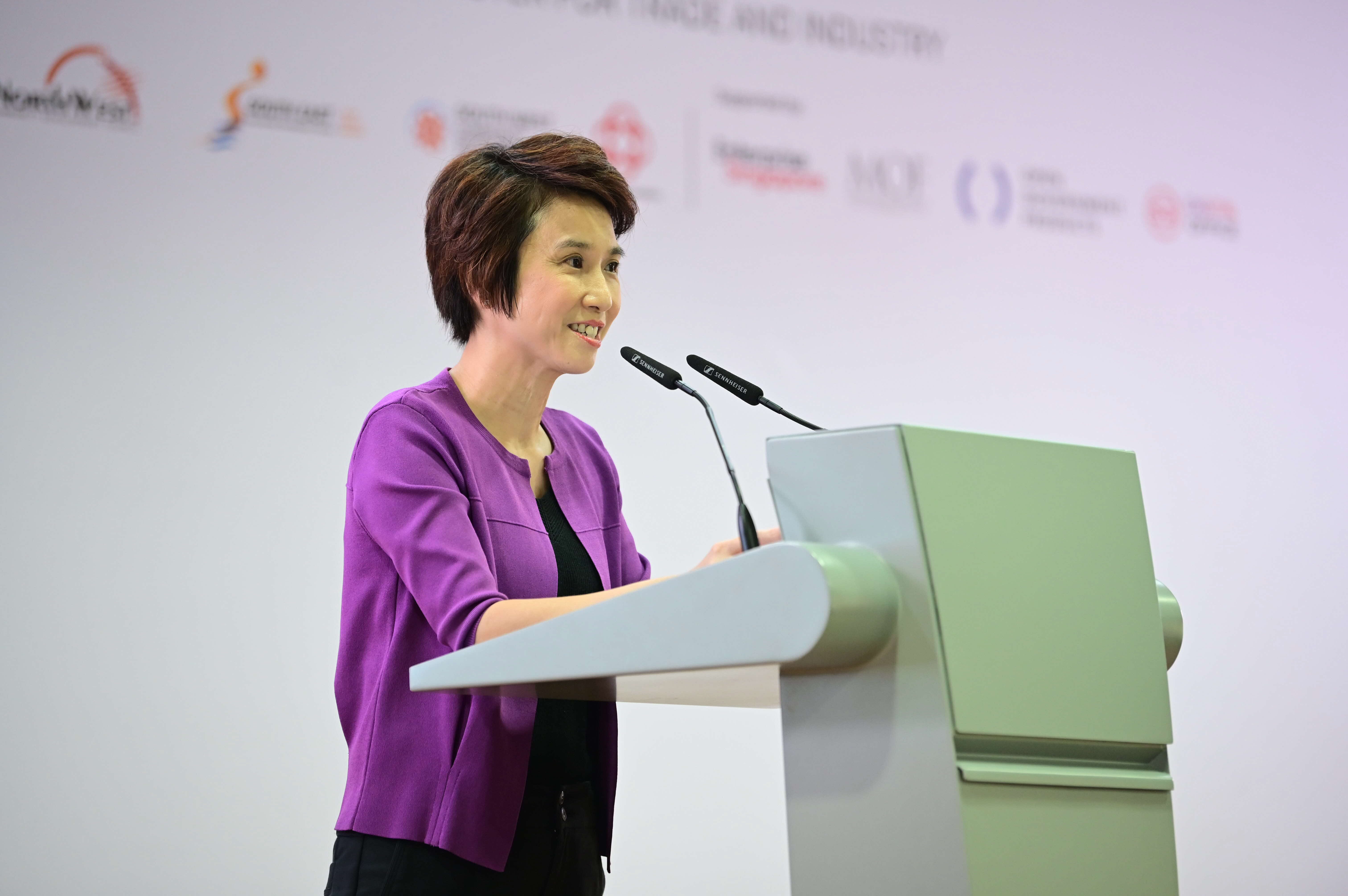 A person in a purple jacket stands at a podium with logos on the backdrop and speaks into two microphones.