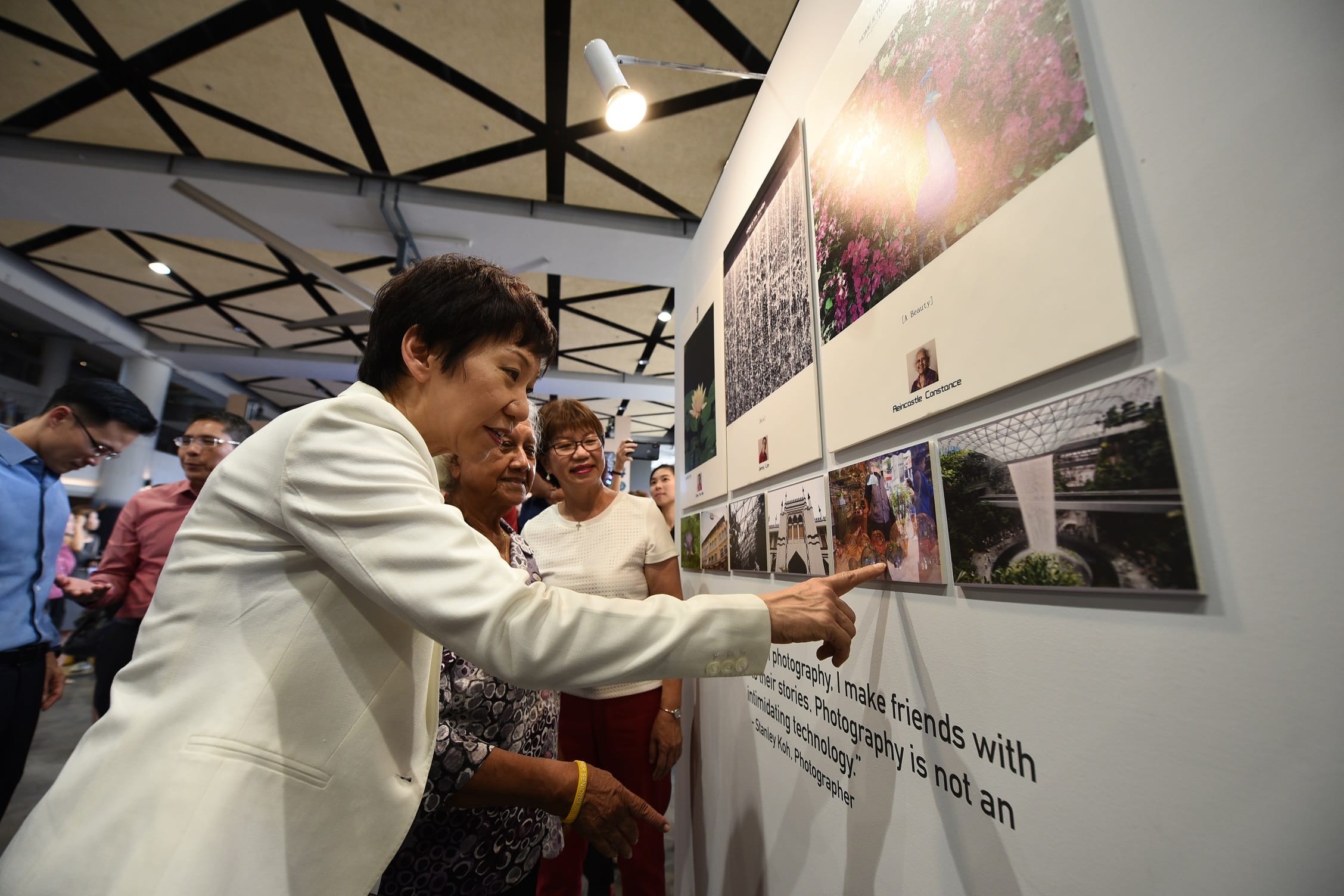 A person in a white jacket points at photos displayed on a gallery wall, surrounded by other blurred individuals.