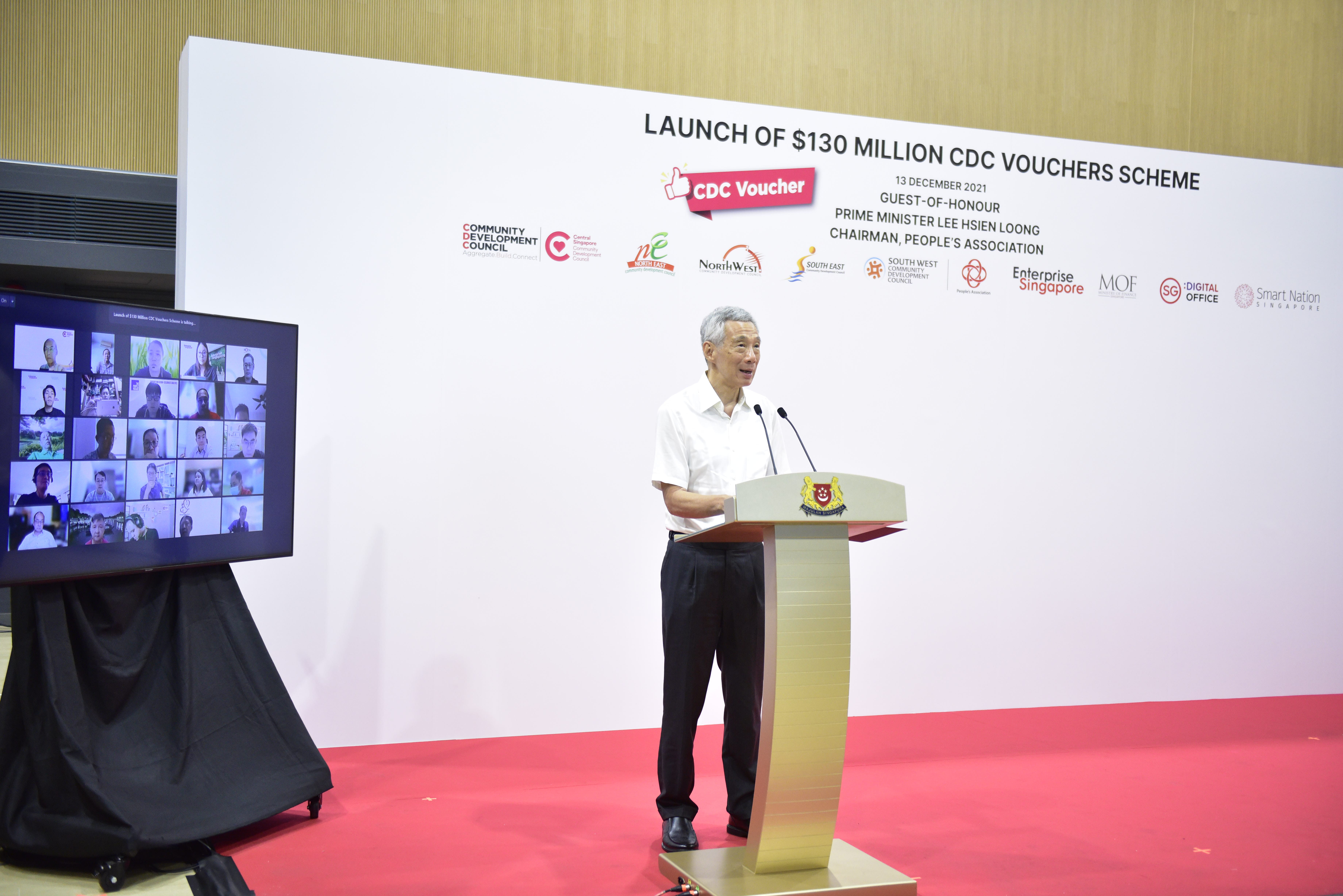 A man speaks at a podium with a red carpet, beside a screen showing participants of a virtual meeting.