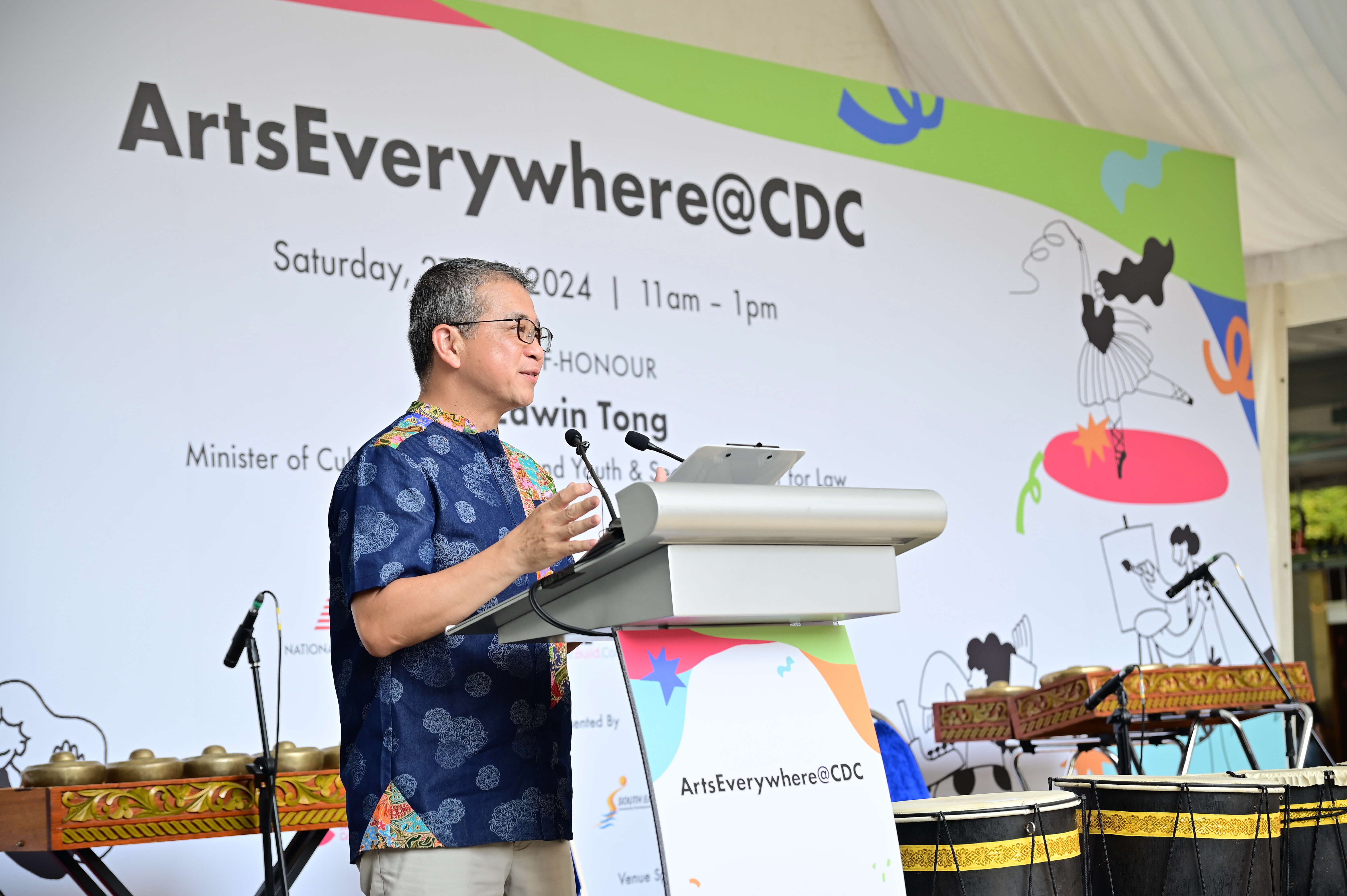 A man stands at a podium speaking at the ArtsEverywhere@CDC event, with traditional musical instruments in the background.