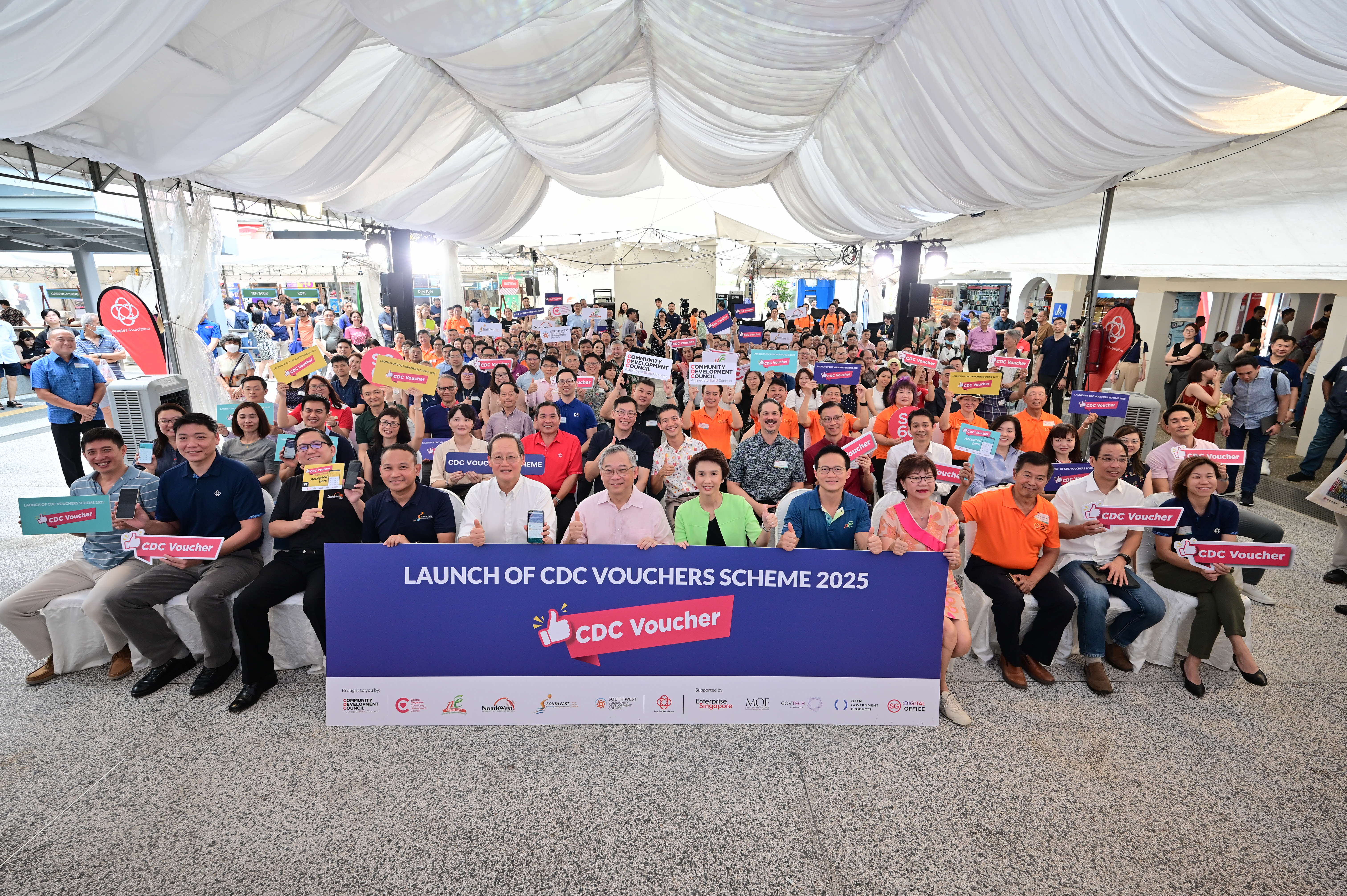 A large group holds colorful signs at the launch event for the CDC Vouchers Scheme 2025 in a decorated space.