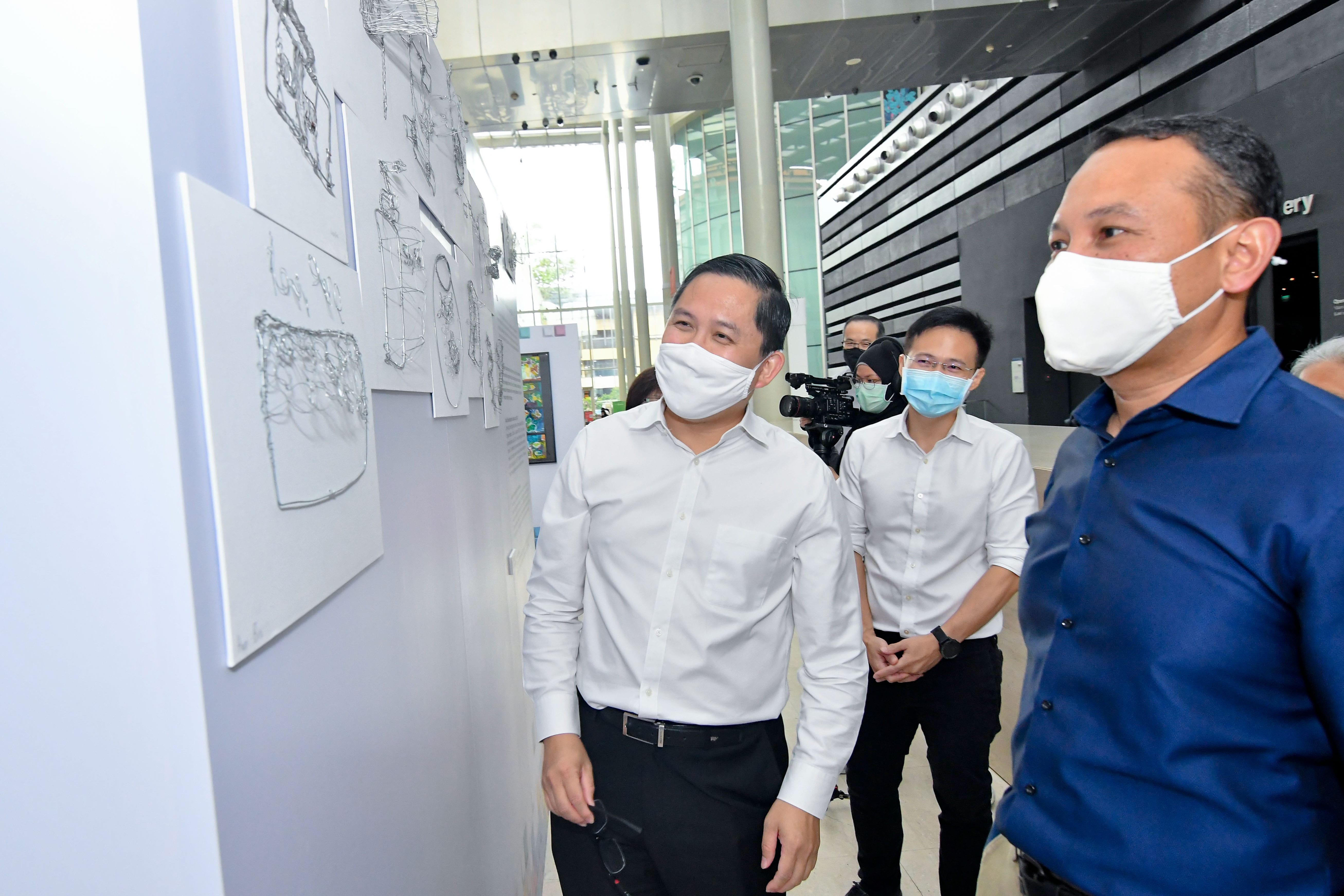 People in white shirts and a blue shirt are standing next to a wall displaying sketches and drawings in a spacious building.