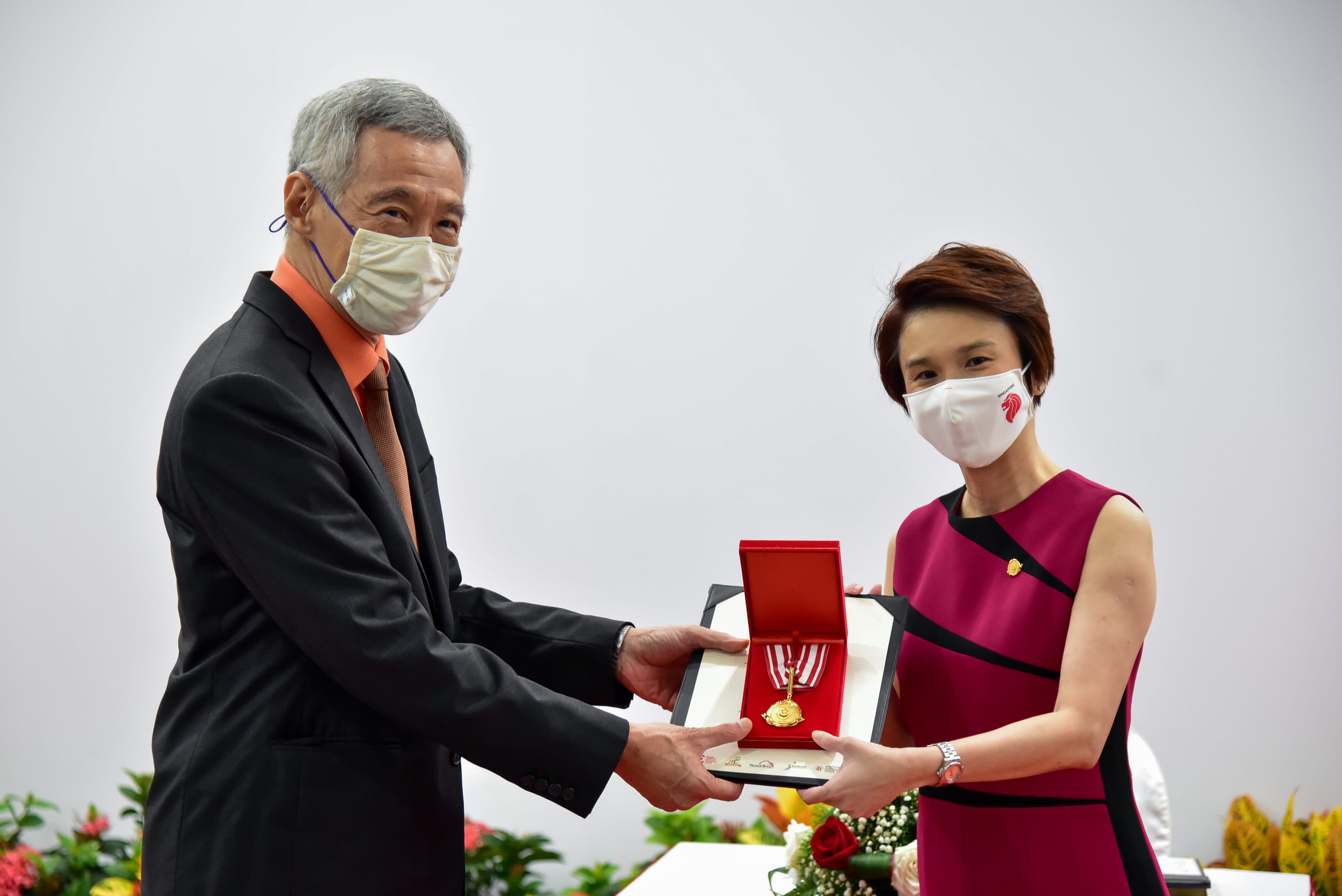 Two individuals exchange a red box with a gold medal inside; one wears a suit, the other a red dress.