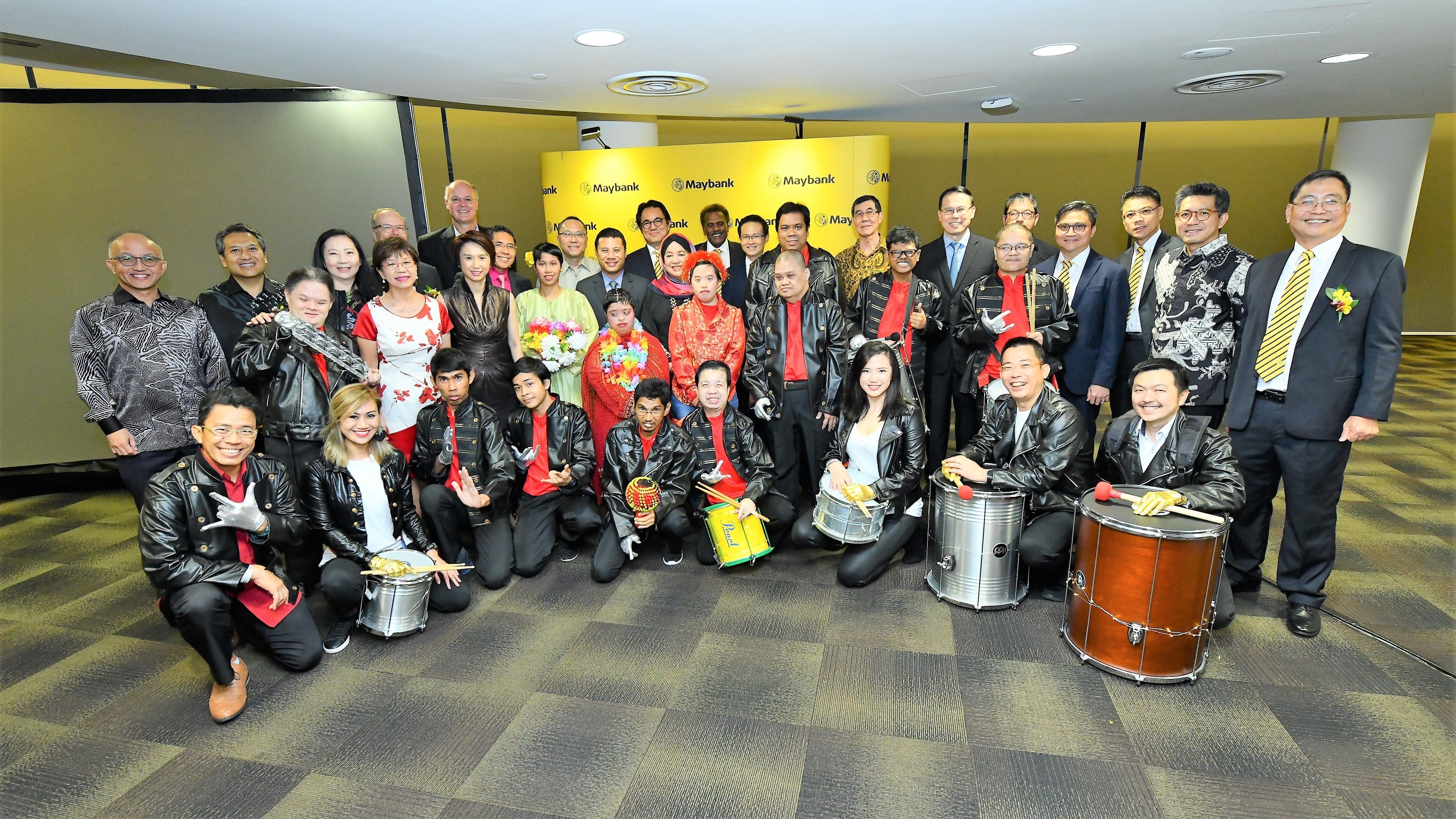 A large group of people are posing together, including some holding drums, in front of a yellow Maybank backdrop.