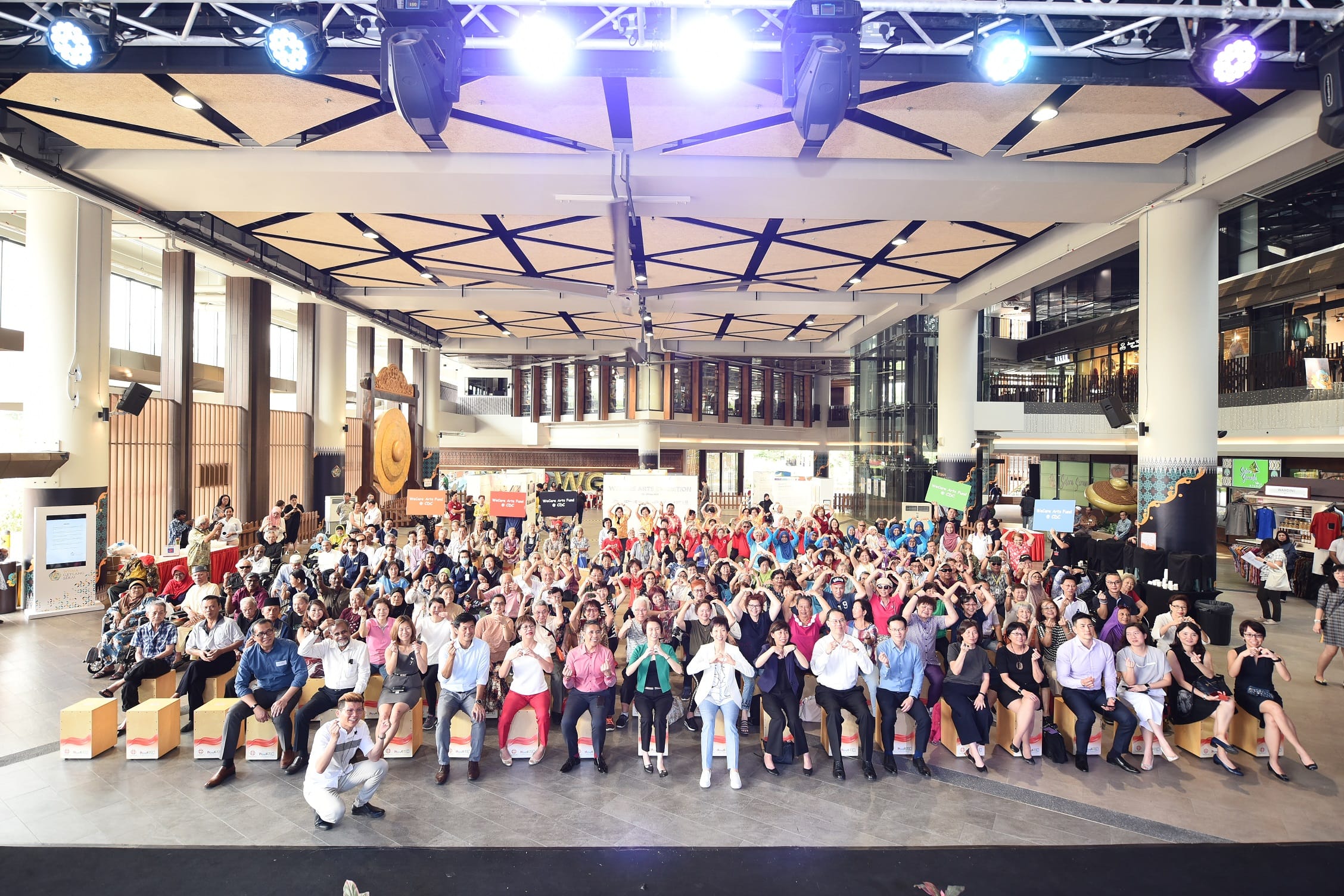 A large group of people is seated in a spacious, brightly lit hall with high ceilings and modern architecture.