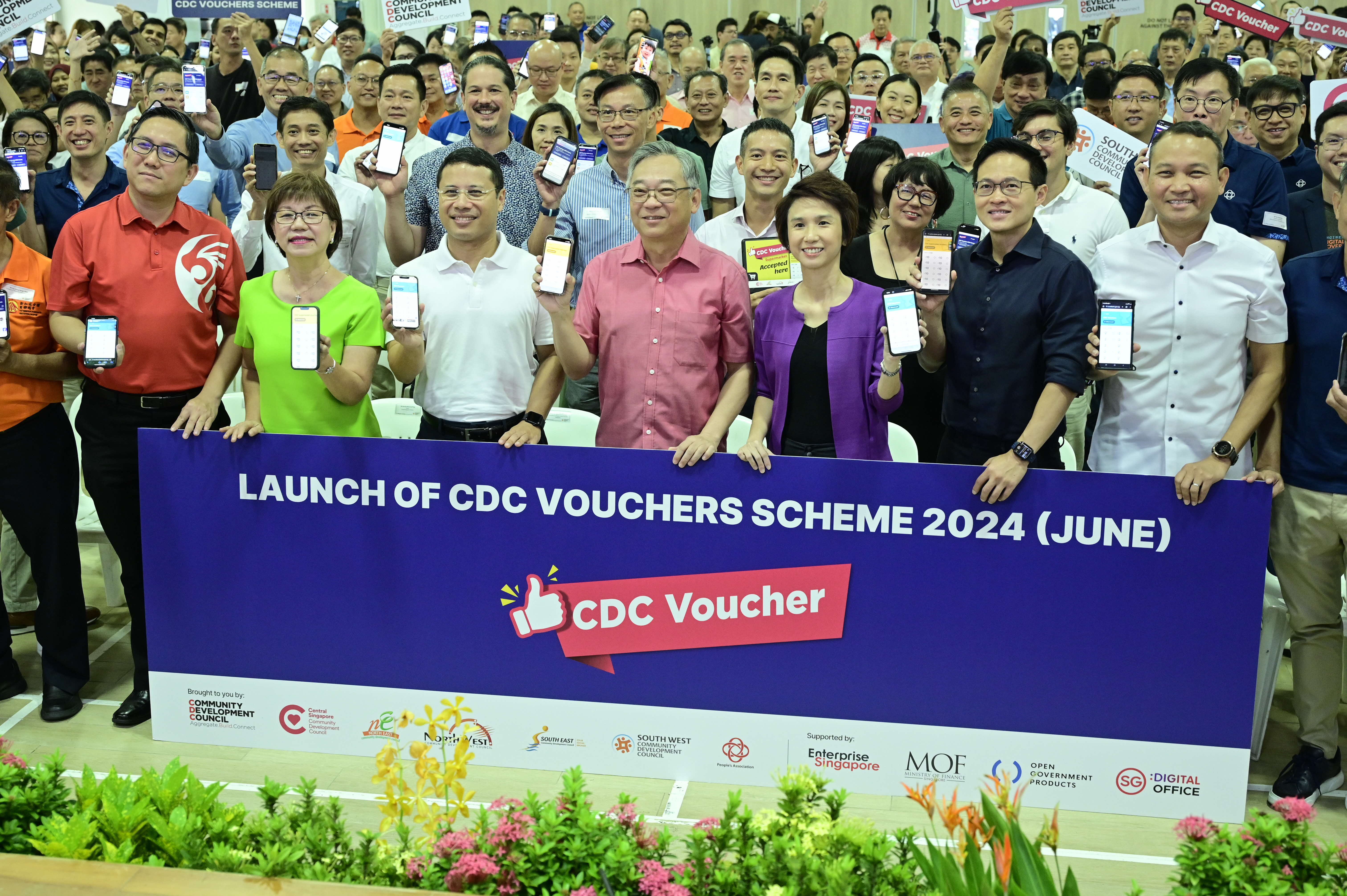 People holding up phones and vouchers stand behind a large sign that reads "Launch of CDC Vouchers Scheme 2024 (June)."