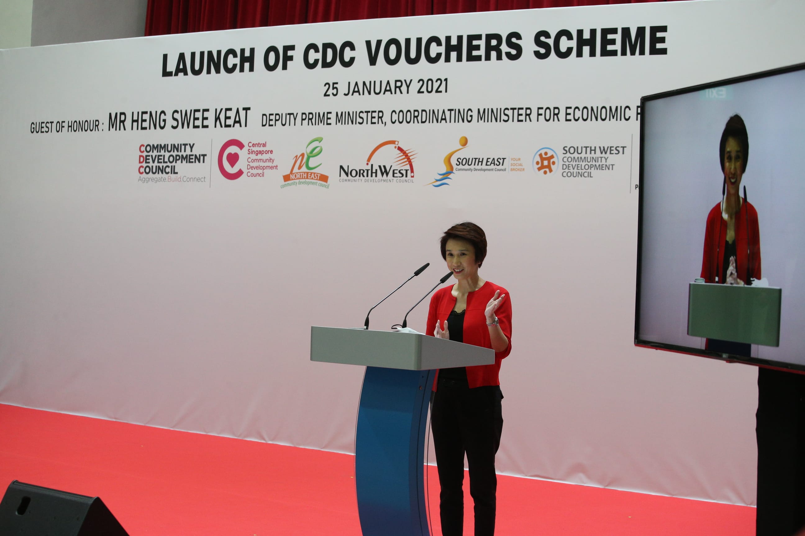 A person in a red jacket speaks at a podium on a stage during the launch of CDC Vouchers Scheme, with a screen beside them.