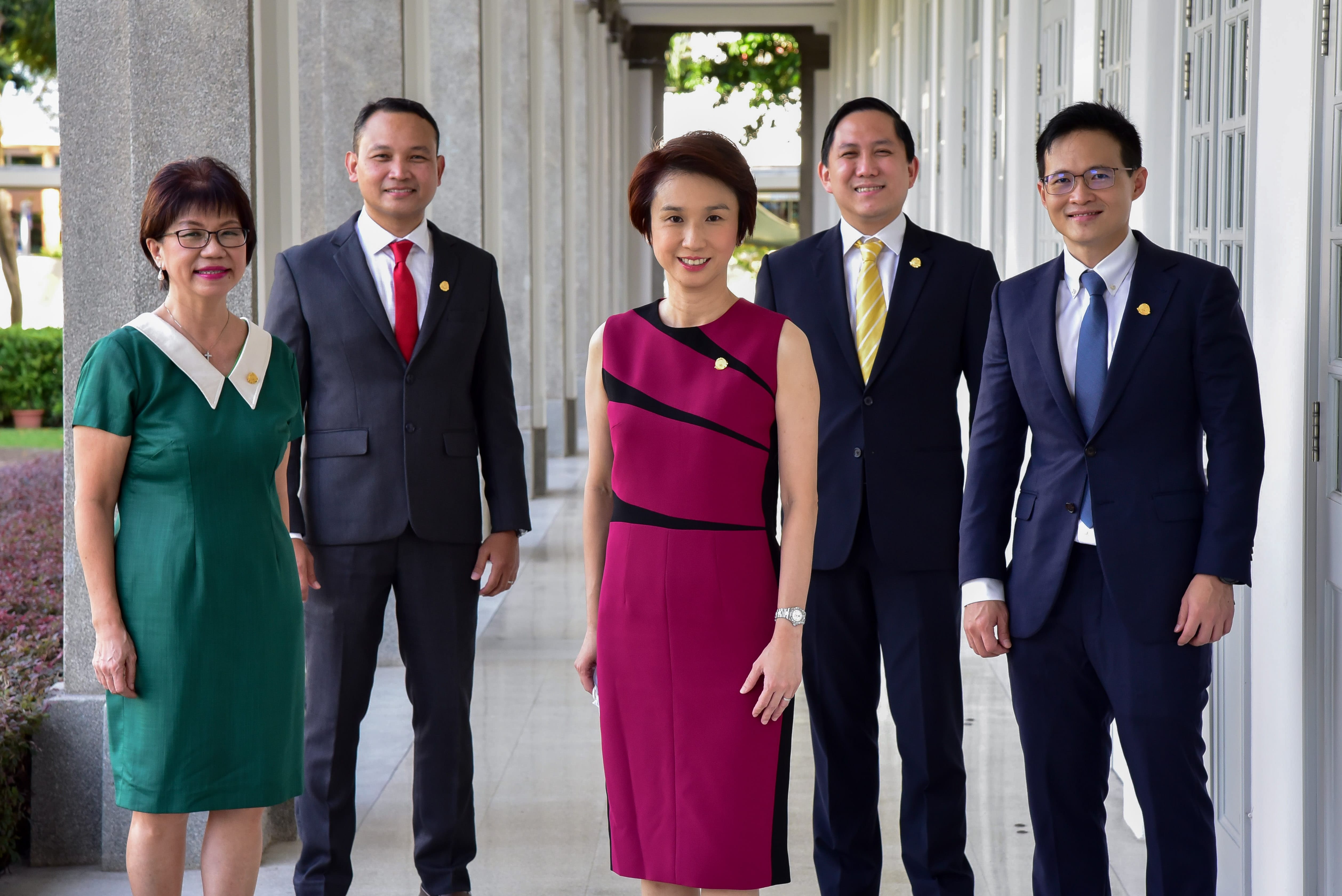 Five people stand in a corridor, with four wearing formal attire and one in a green dress.