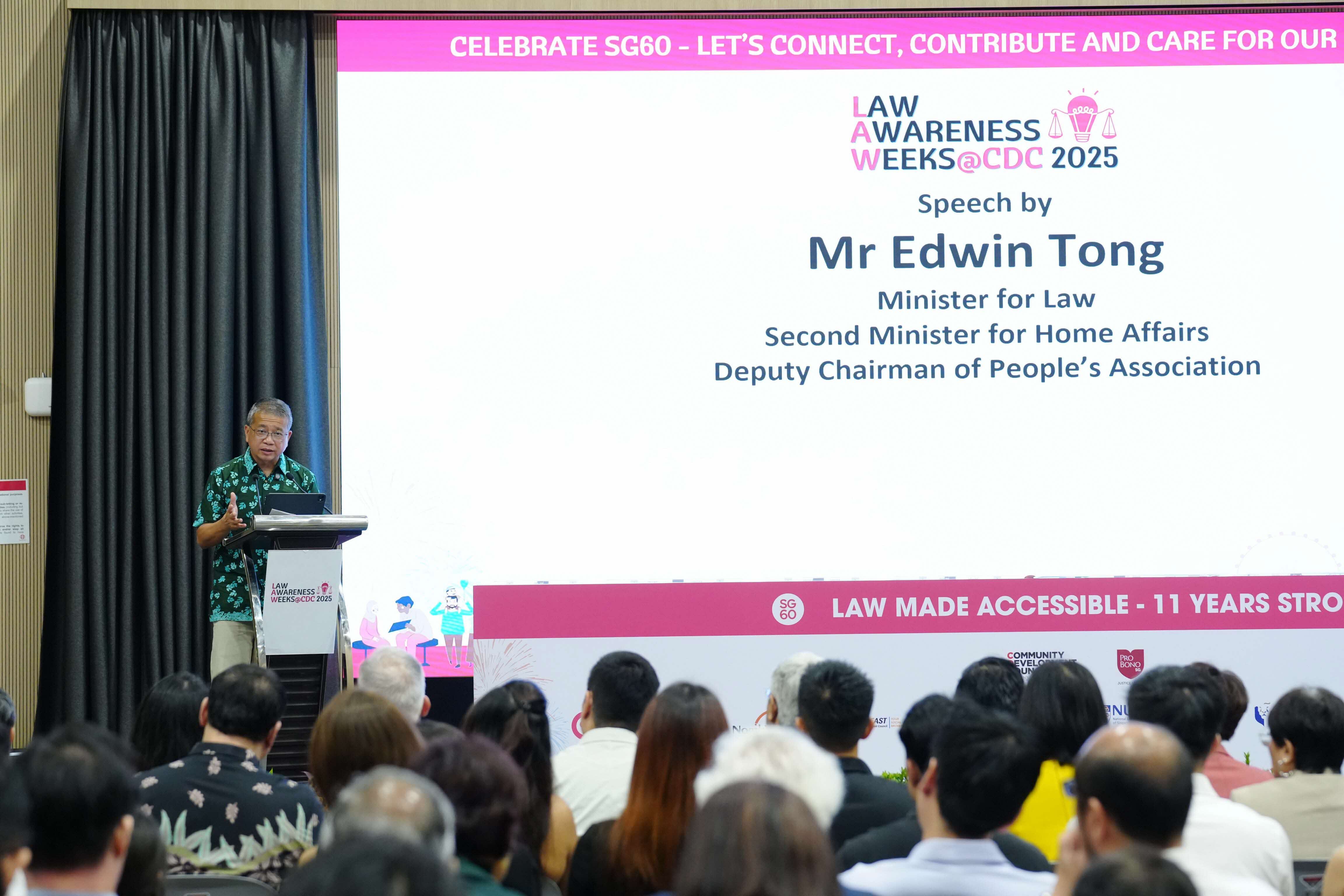 A speaker at a podium addresses an audience, with a large screen displaying details of Mr. Edwin Tong's speech.