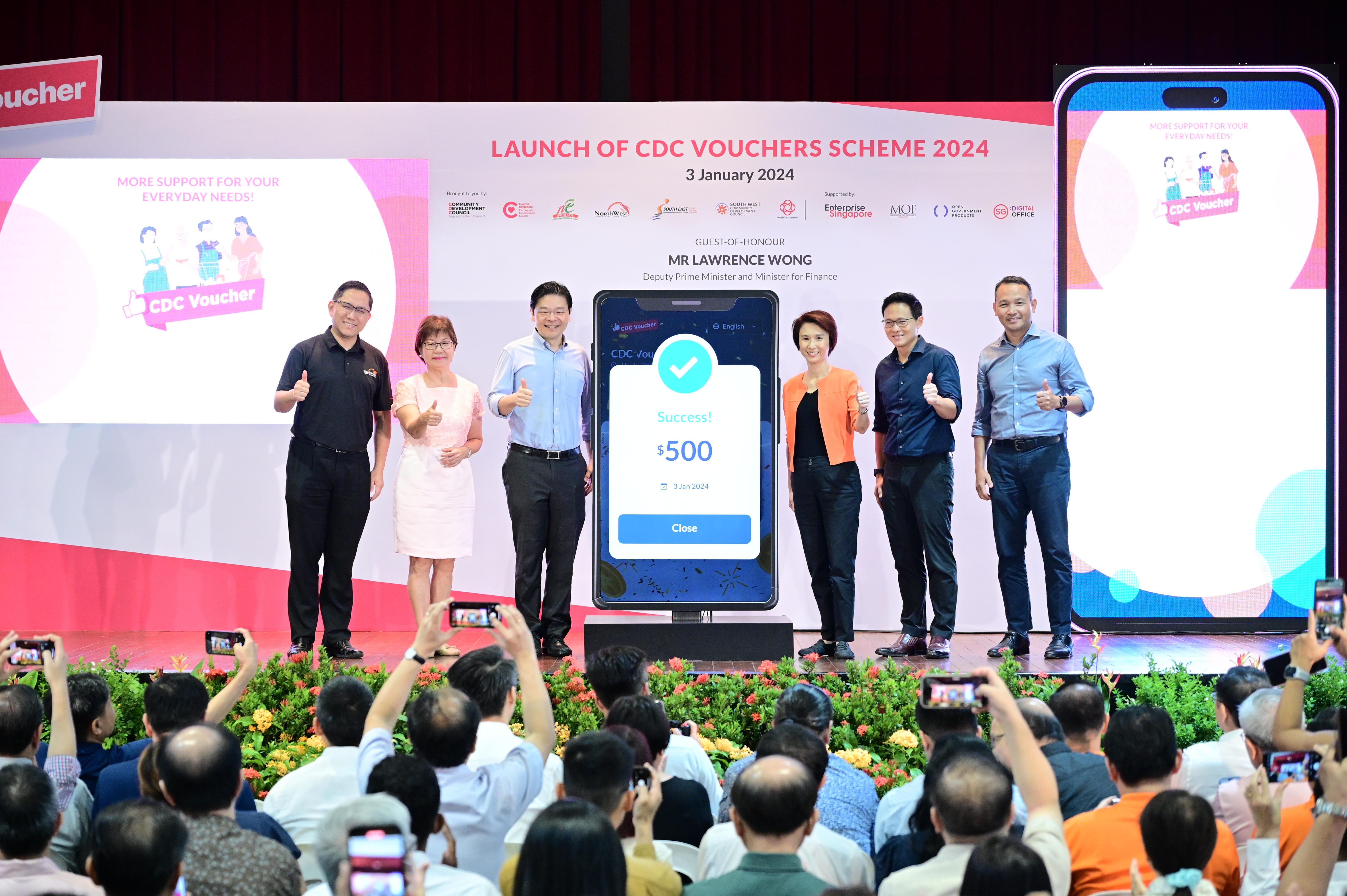 Several people on stage at the launch of CDC Vouchers Scheme 2024, with a large smartphone display showing a $500 success message.