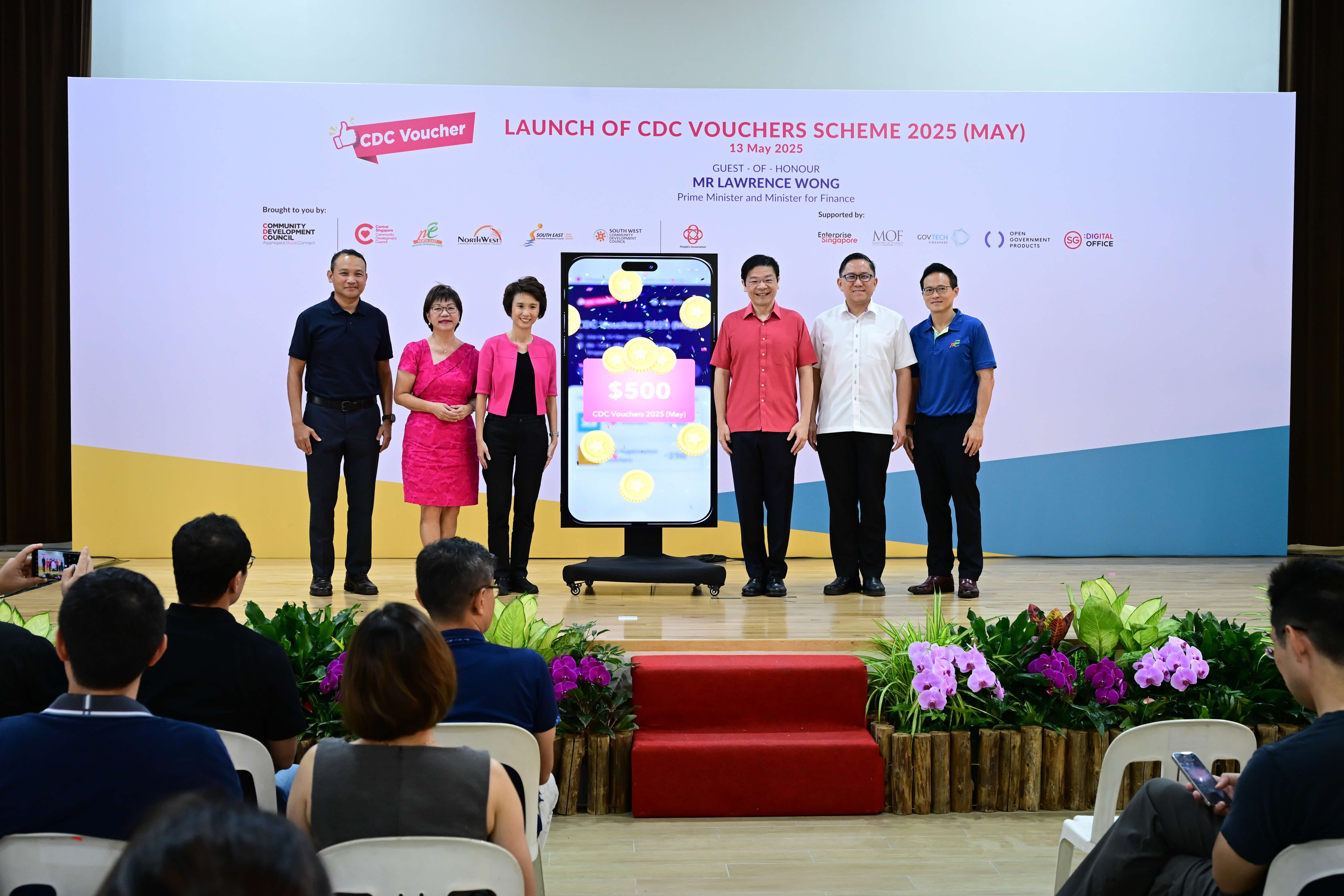 Five people stand on stage near a large smartphone displaying "$500 CDC Vouchers Scheme 2025" at an event.