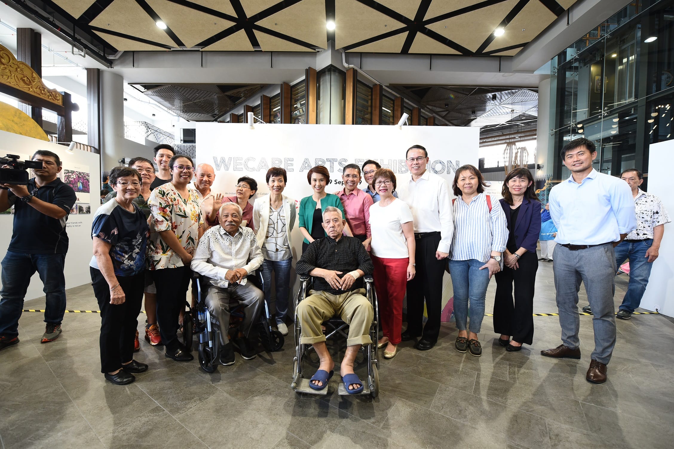 A group of people, including two in wheelchairs, stand in front of a "WE CARE ARTS EXHIBITION" sign in an indoor space.