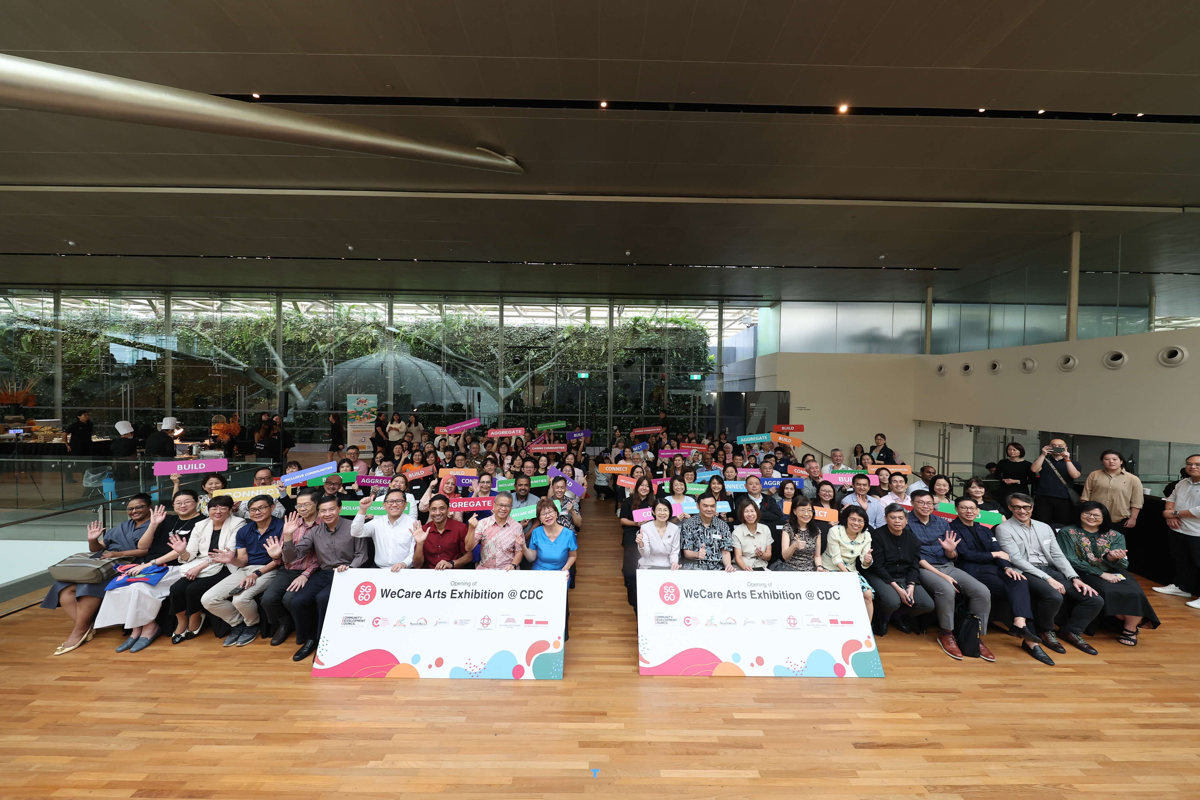 Group photo at an indoor event, with attendees holding colorful signs, seated behind banners reading "WeCare Arts Exhibition @ CDC."