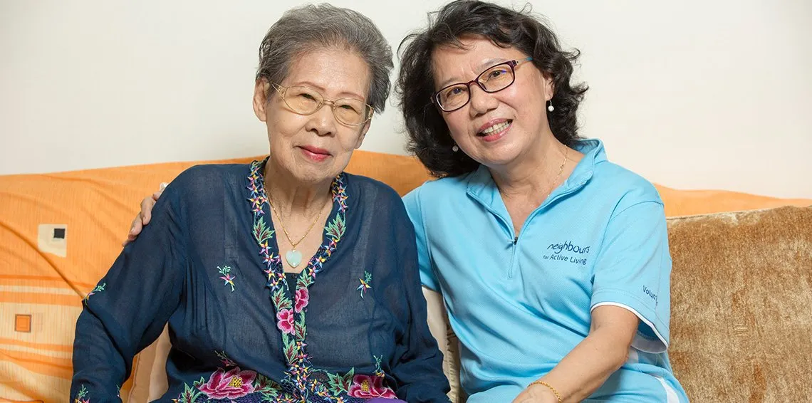 An elderly woman in a navy blue embroidered blouse and a middle-aged volunteer in a light blue polo shirt sitting together on an orange couch, both wearing glasses and smiling at the camera, representing intergenerational bonding through community volunteer work.