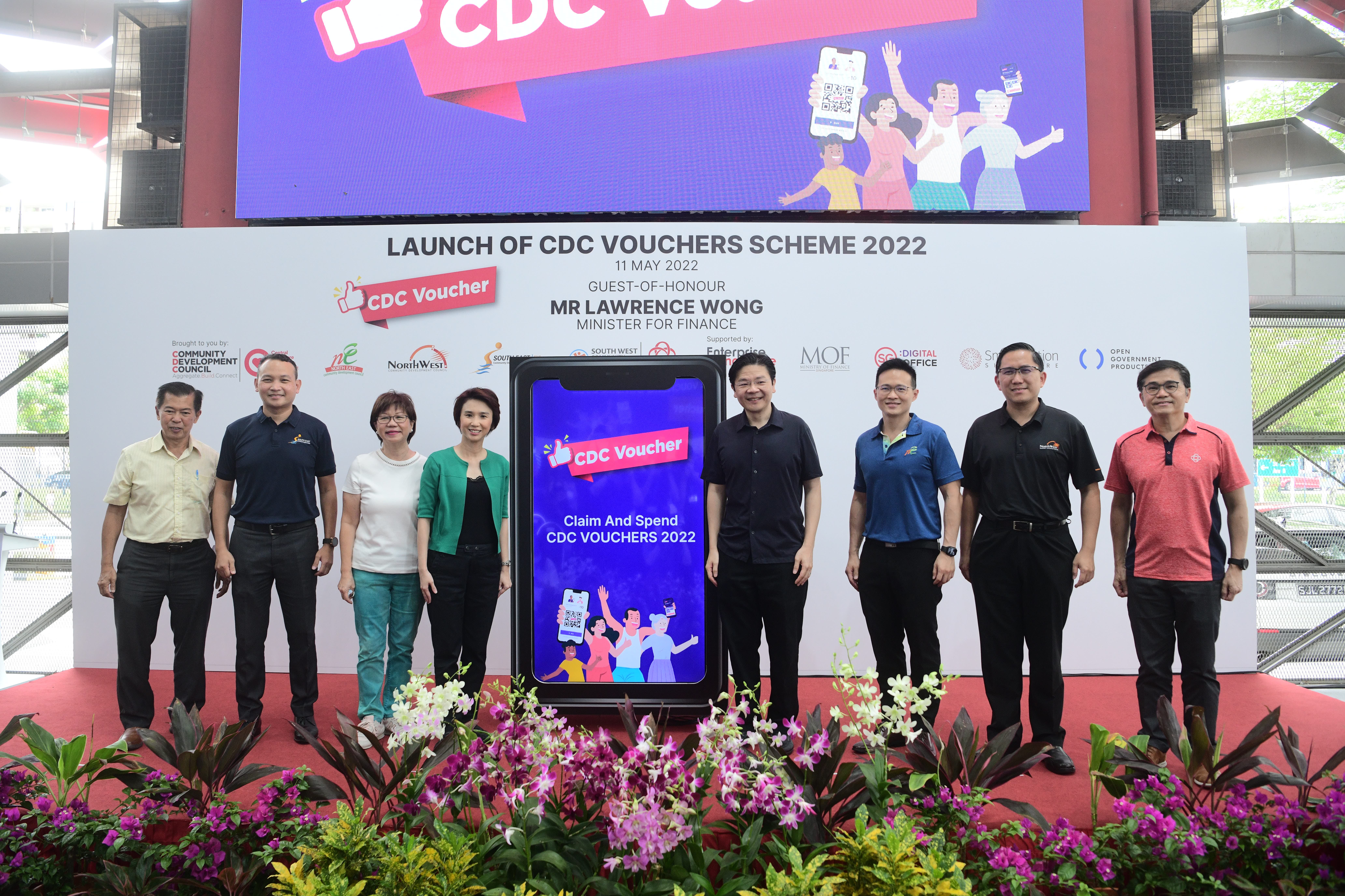 A group of seven people stands on stage with a large sign reading "CDC Voucher" and flowers in front.