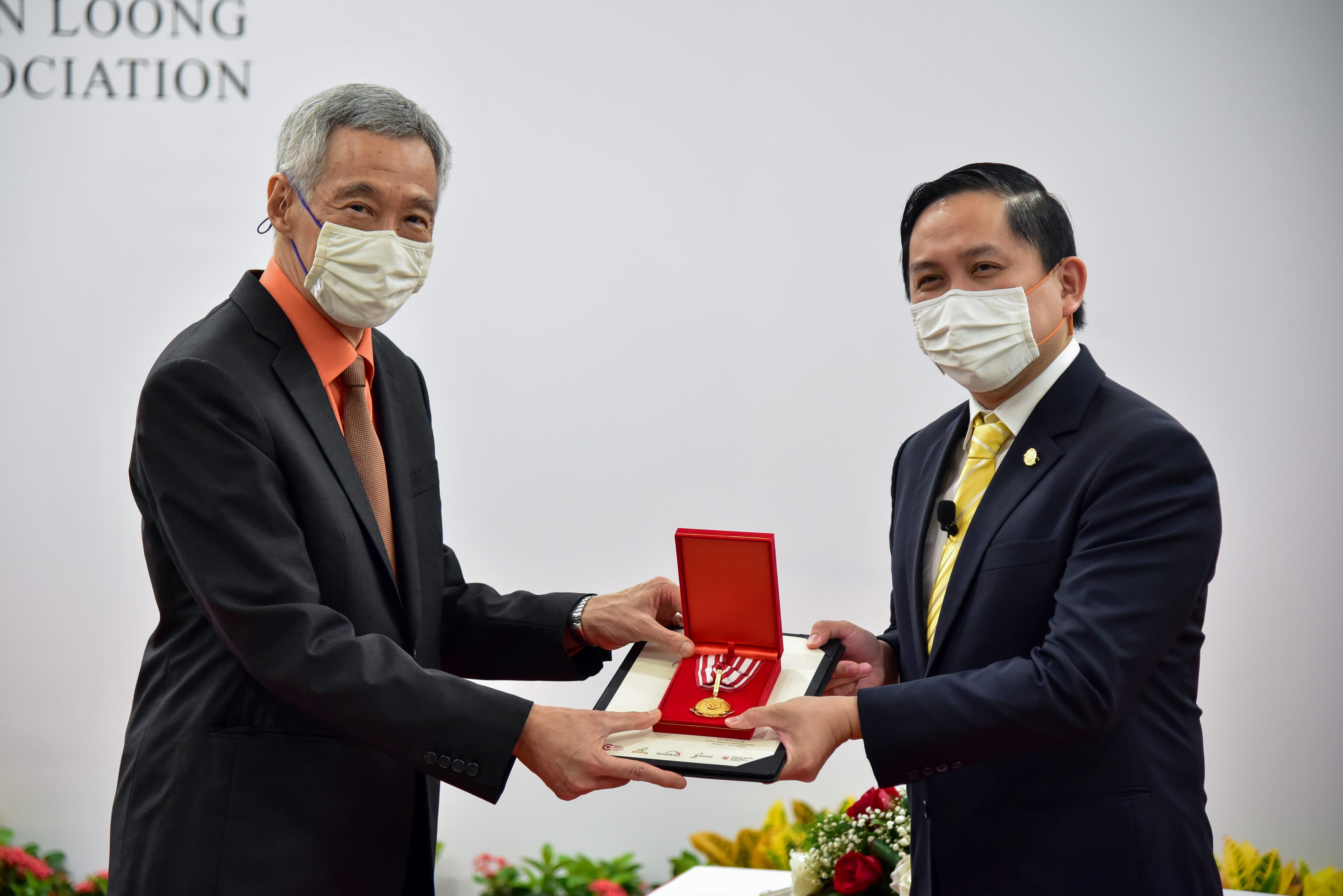 Two people in suits exchange a medal in a red box at an event, with flowers on a table nearby.